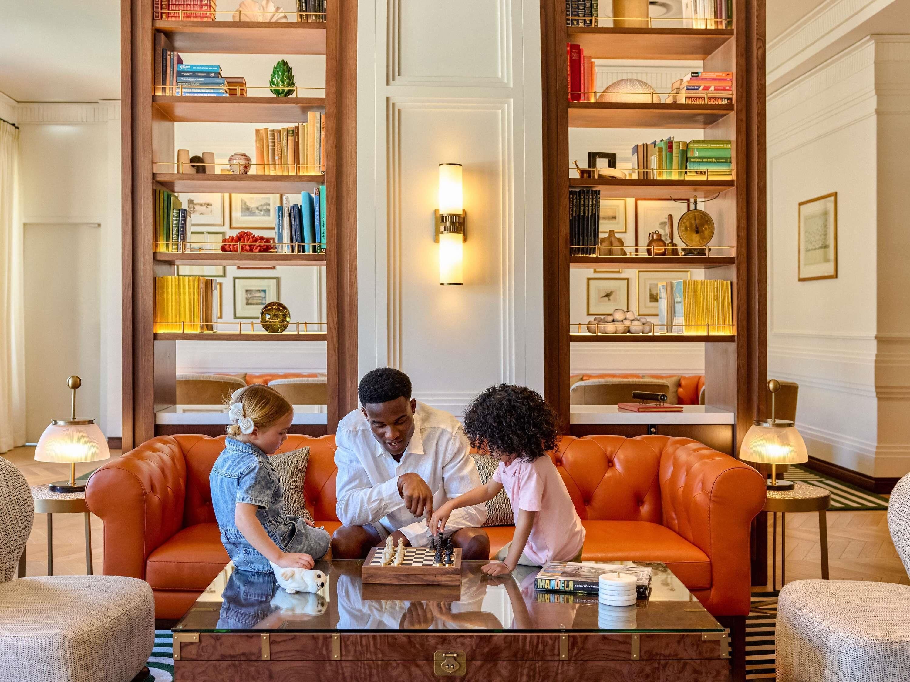 A cozy living room with a leather sofa, a coffee table, and a chessboard. There are bookshelves with books and decorative items.