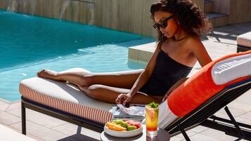 A person relaxing by a poolside lounge chair with a drink and a bowl of fruit.