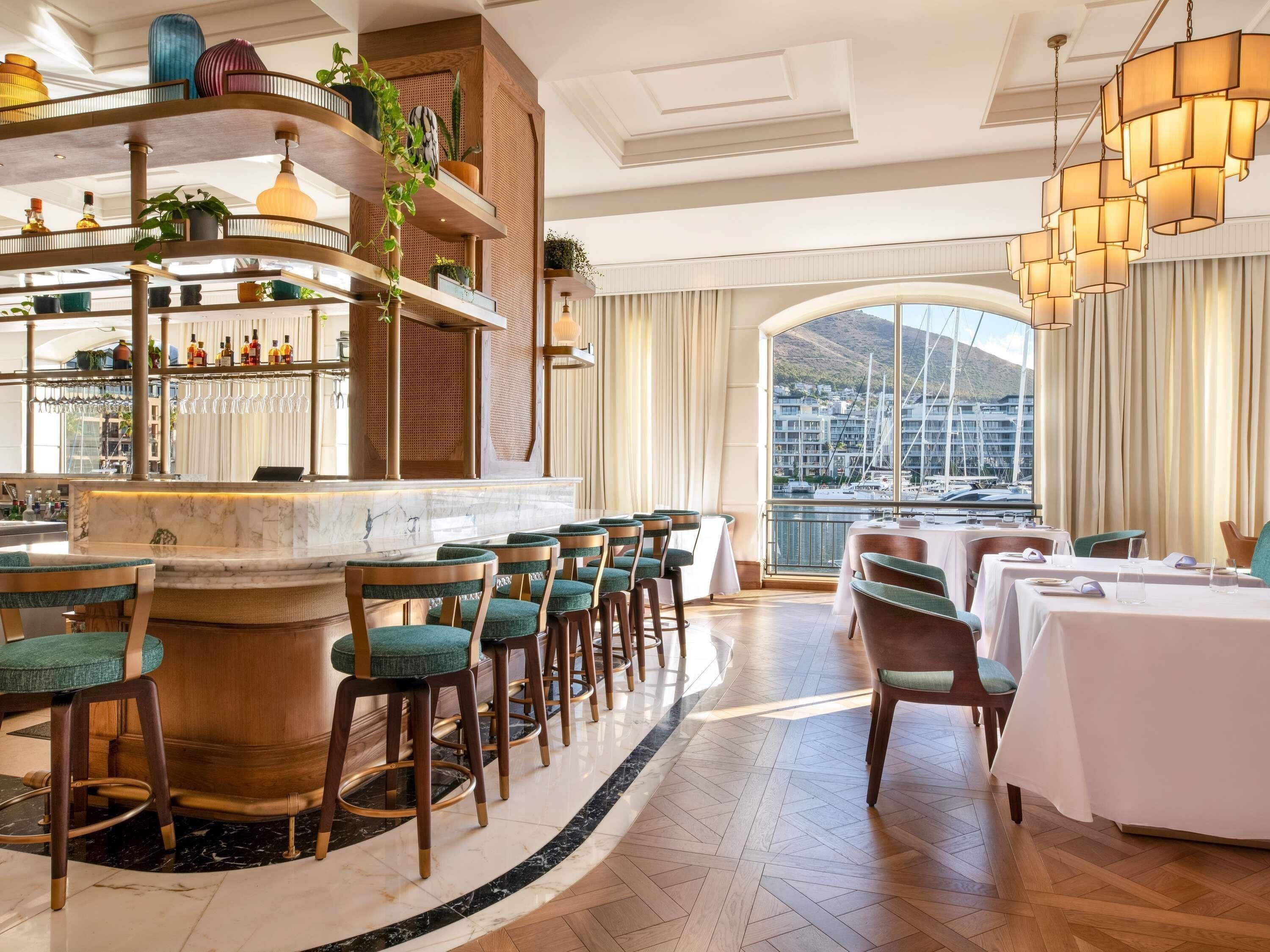 Un espace bar avec des tabourets hauts et un comptoir en marbre, un espace repas avec des tables et des chaises rondes, et une grande fenêtre offrant une vue sur les bateaux et les bâtiments.