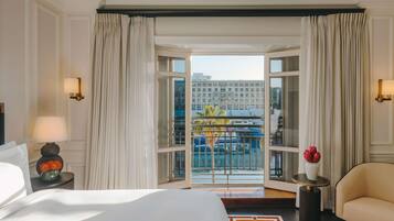 A hotel room with a large bed, a sitting area with a chair, a small table with a vase of flowers, and a balcony with a view of buildings and a blue boat.