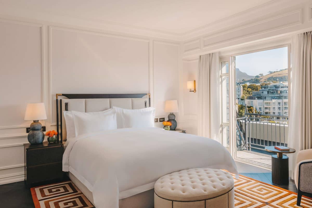 Habitación de hotel con una cama grande, un reposapiés redondo, vistas a edificios y colinas a través de la ventana, y un balcón con barandilla.