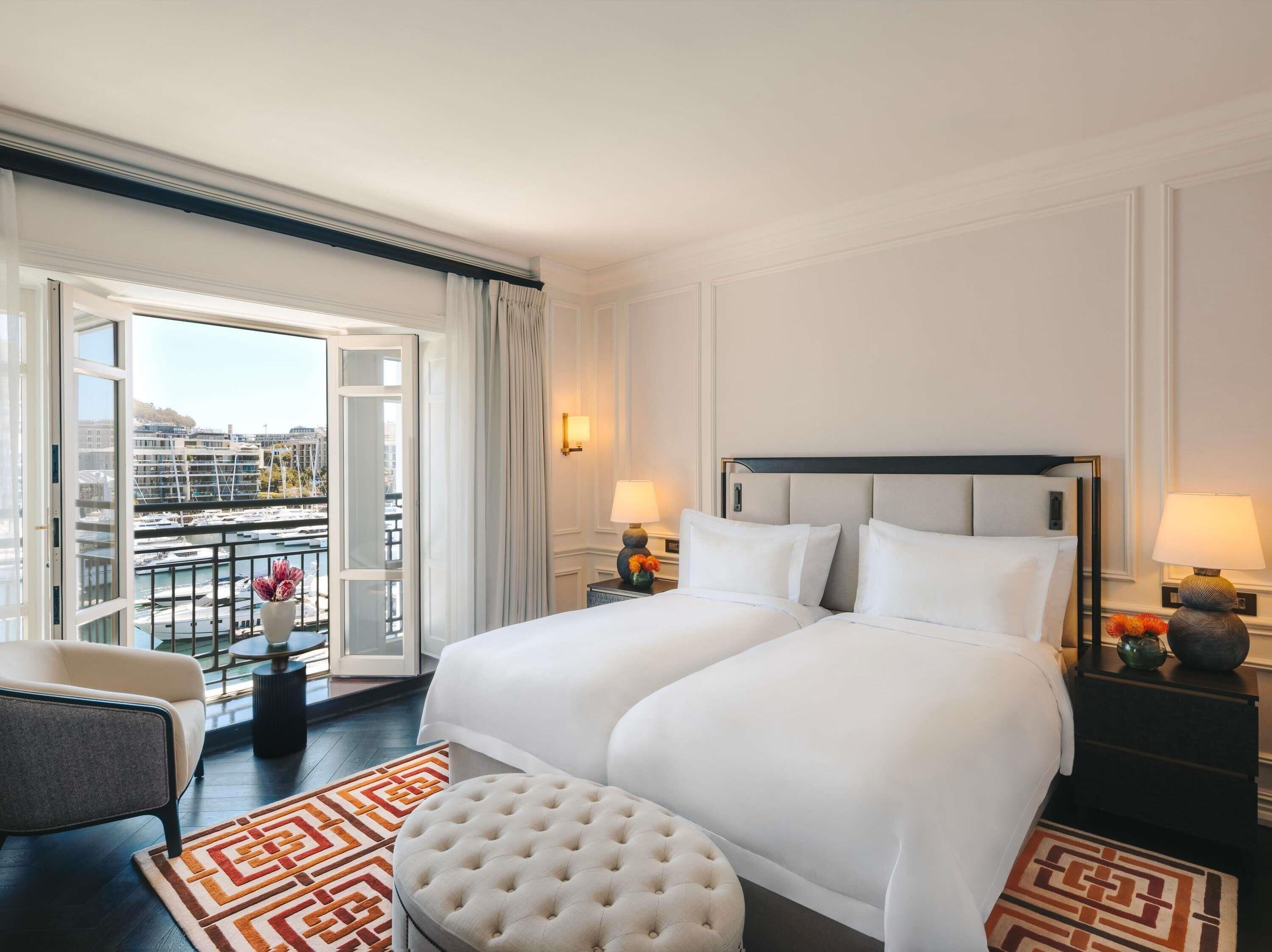 A hotel room with a large bed, two bedside tables, a chair, a view of the city through a balcony, and a patterned rug.