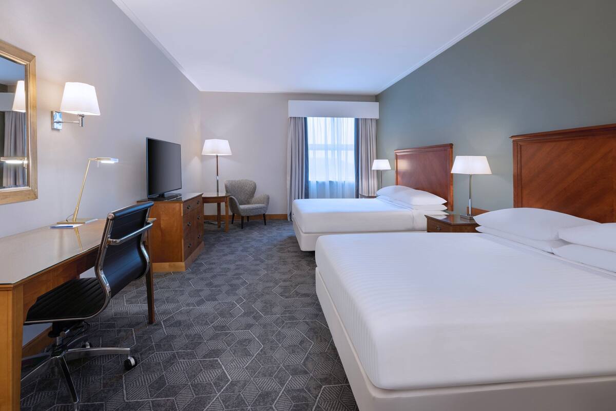 A hotel room with two beds, a desk with a chair, a TV, and a small seating area with a chair and table.