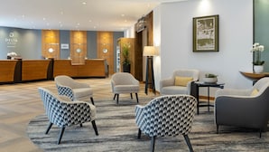 A hotel reception area with a wooden reception desk, several armchairs, a small table with a lamp, and a framed picture on the wall. - Delta Hotels Bexleyheath (Bexleyheath)