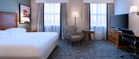 Ein Hotelzimmer mit einem großen Bett, einem Schreibtisch mit Stuhl, einem Fernseher und einem kleinen Tisch.