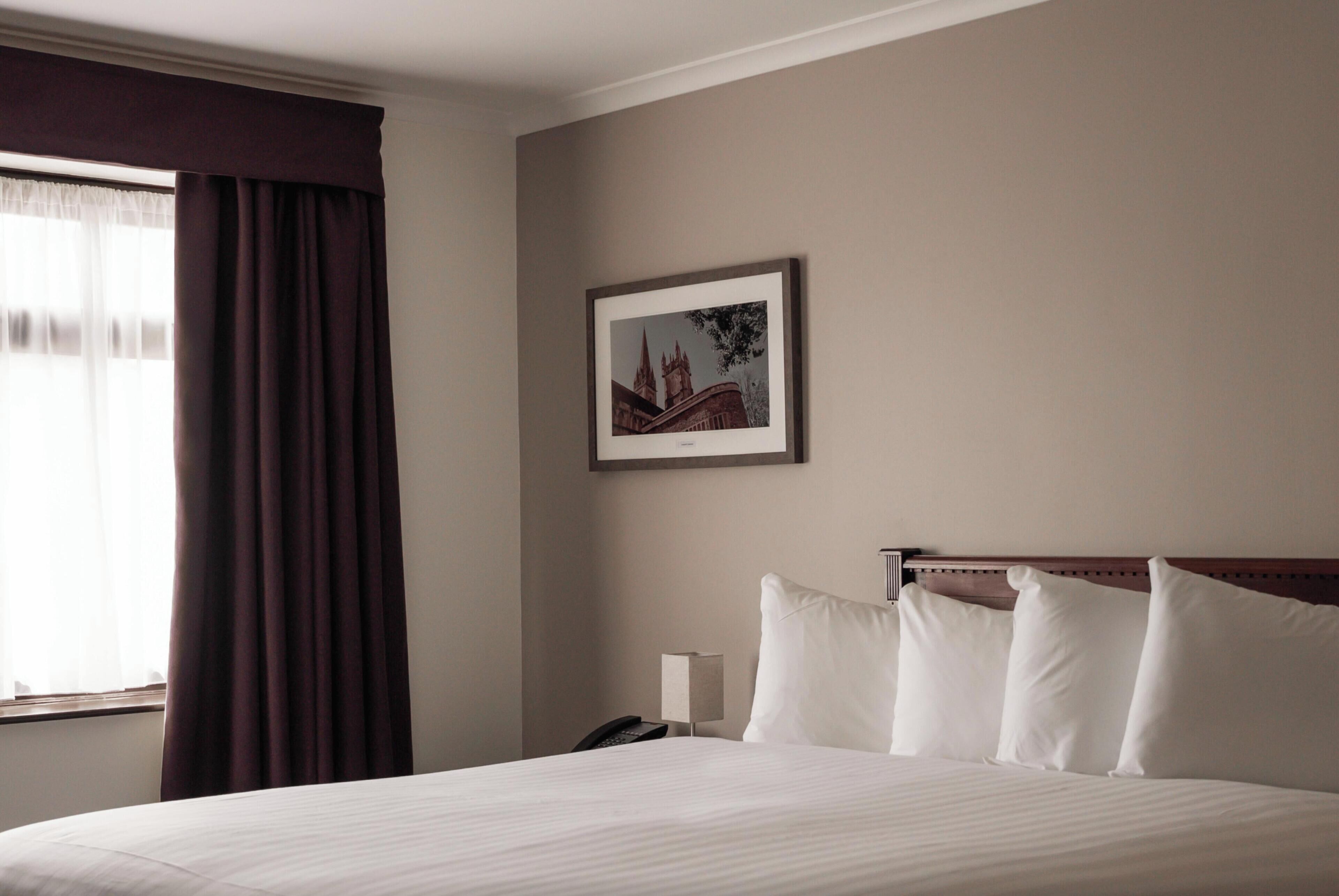 Ein Hotelzimmer mit einem Bett, einem Nachttisch mit Lampe, einem gerahmten Bild an der Wand und einem Fenster mit Vorhängen.
