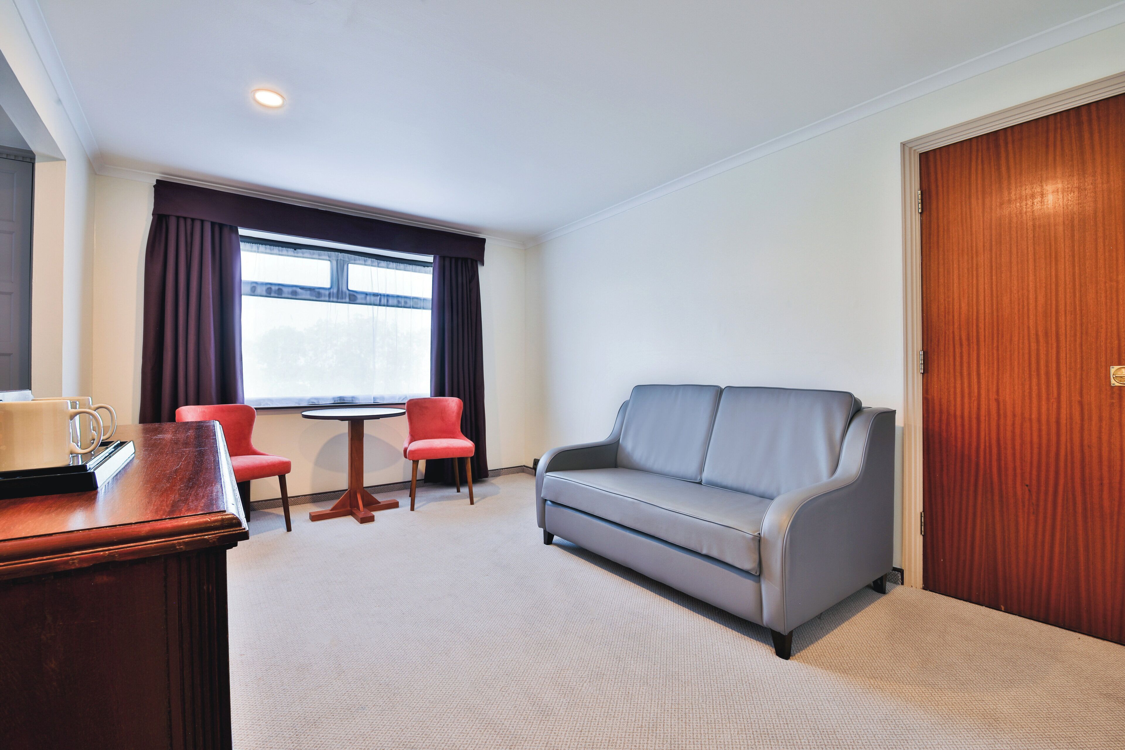 Ein Hotelzimmer mit grauem Sofa, einem roten Sessel, einem Holztisch und einem Fenster mit Vorhängen.