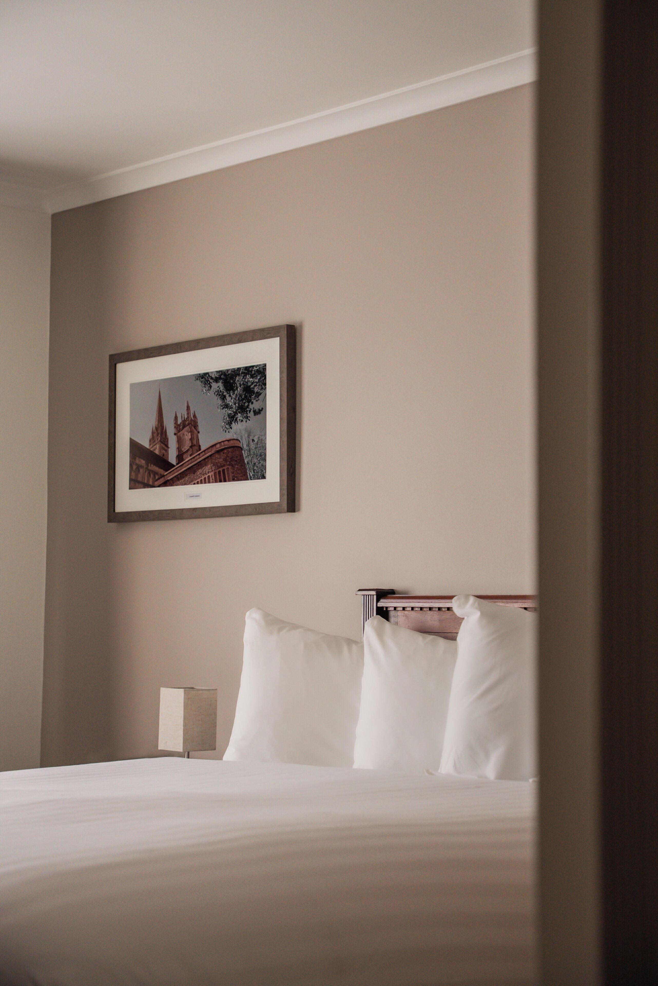 A neatly made bed with white linens, a framed picture of a building on the wall, and a small bedside table with a lamp.
