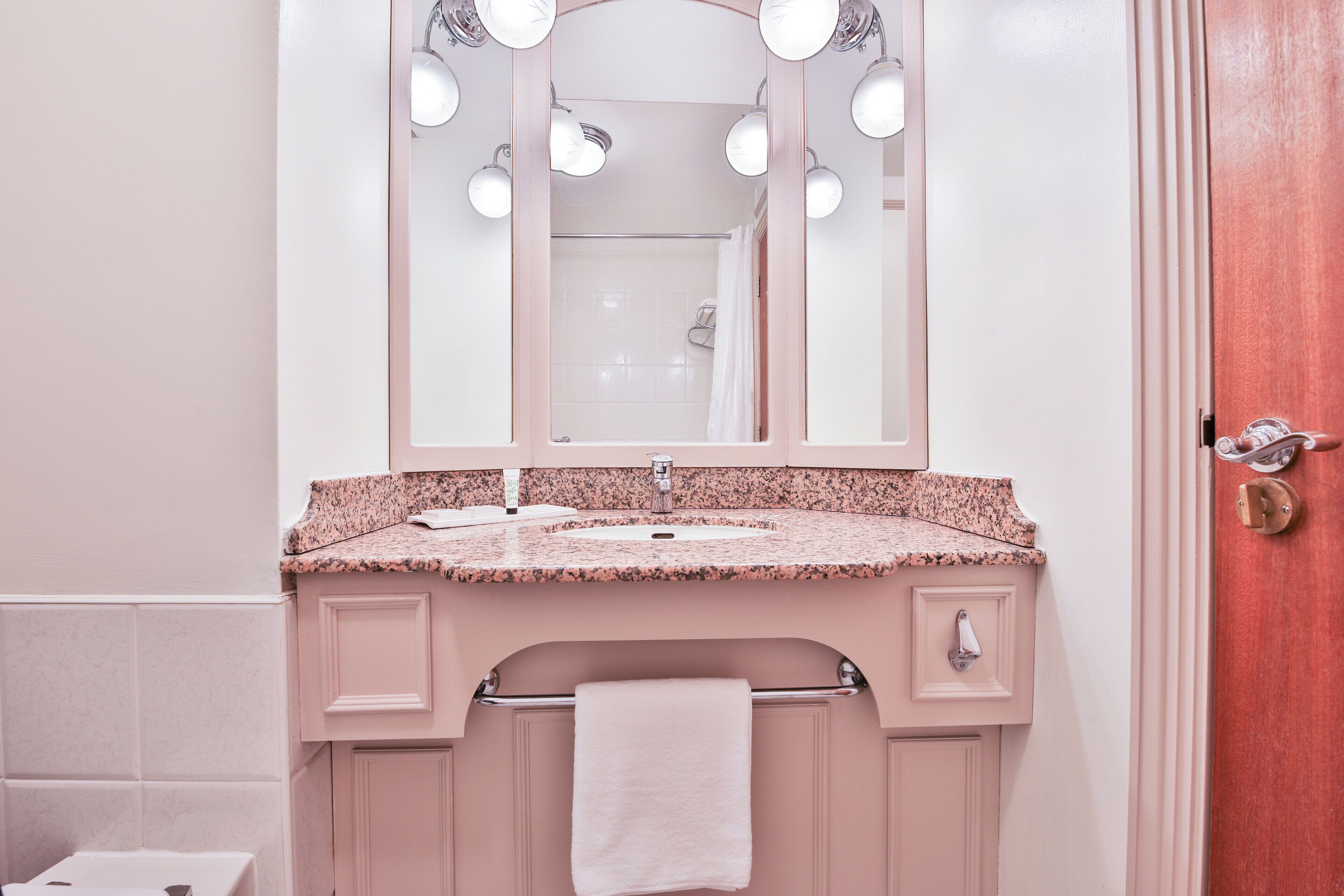 A bathroom with a pink vanity, a mirror with lights, a towel rack, and a door.