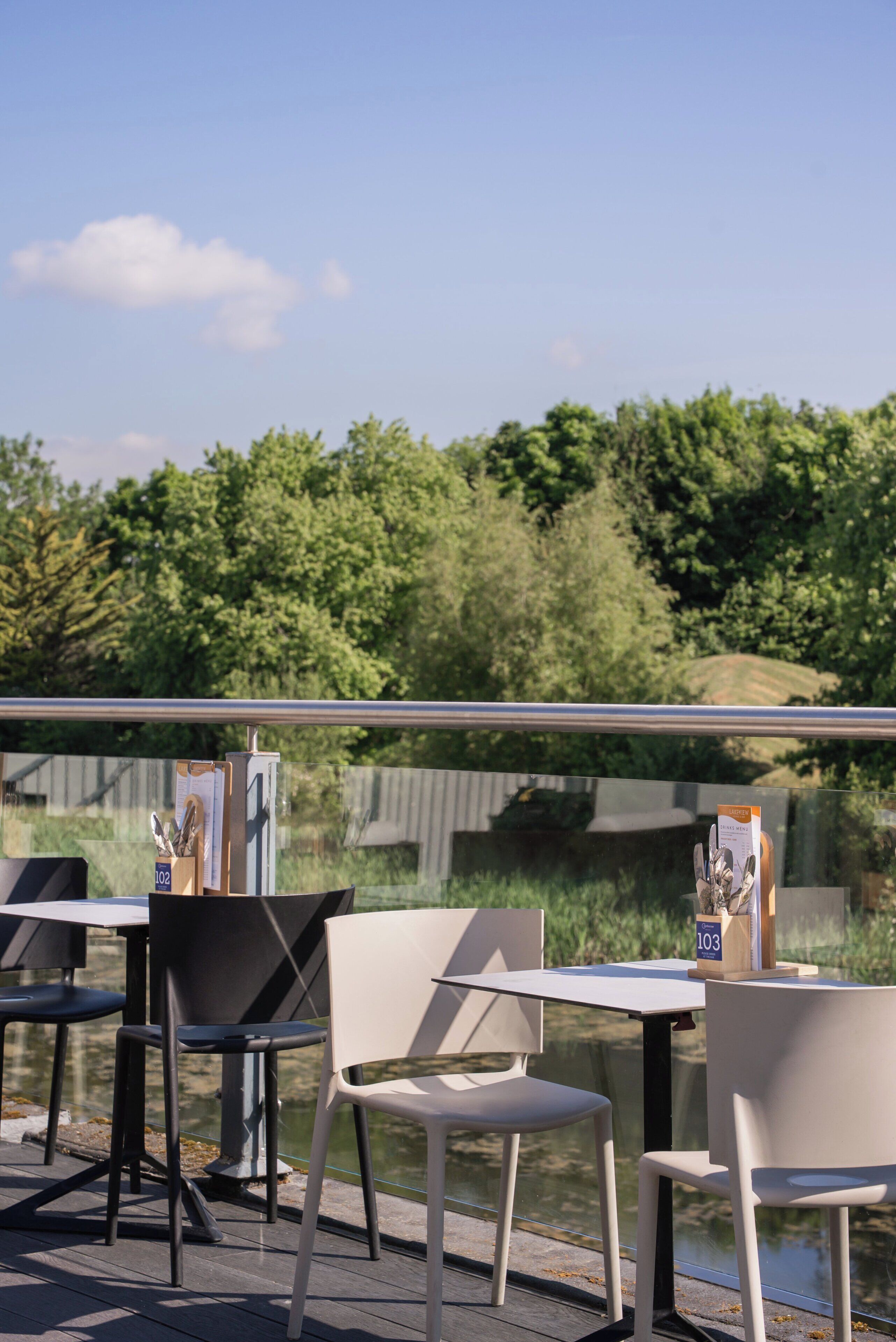 Buiten zitgedeelte met tafels en stoelen, een leuning en uitzicht op water en groen op de achtergrond.