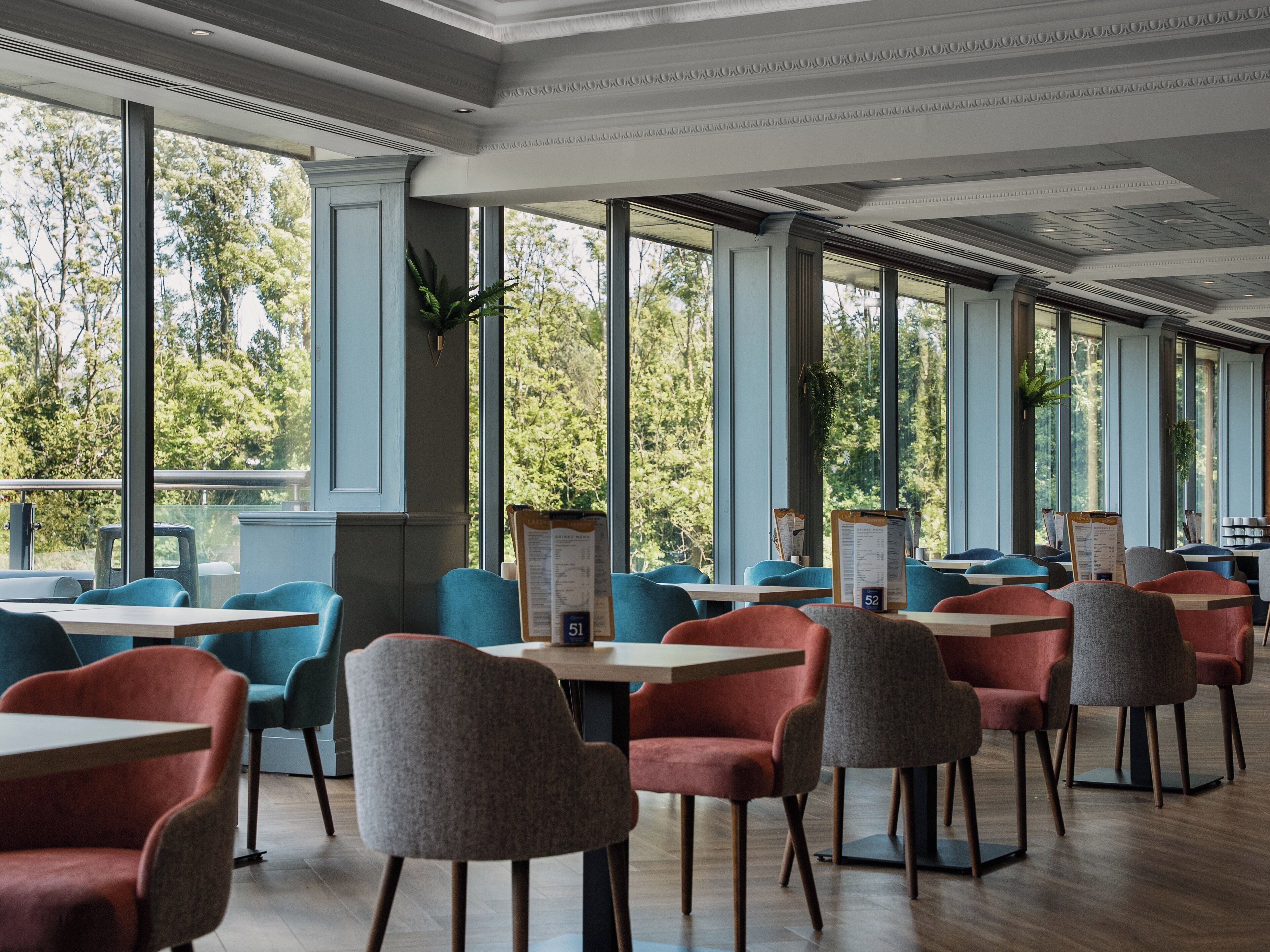 Ein geräumiges Restaurant mit hohen Decken und großen Fenstern, das Blick auf viel Grün bietet. Tische und Stühle sind zum Essen angeordnet.