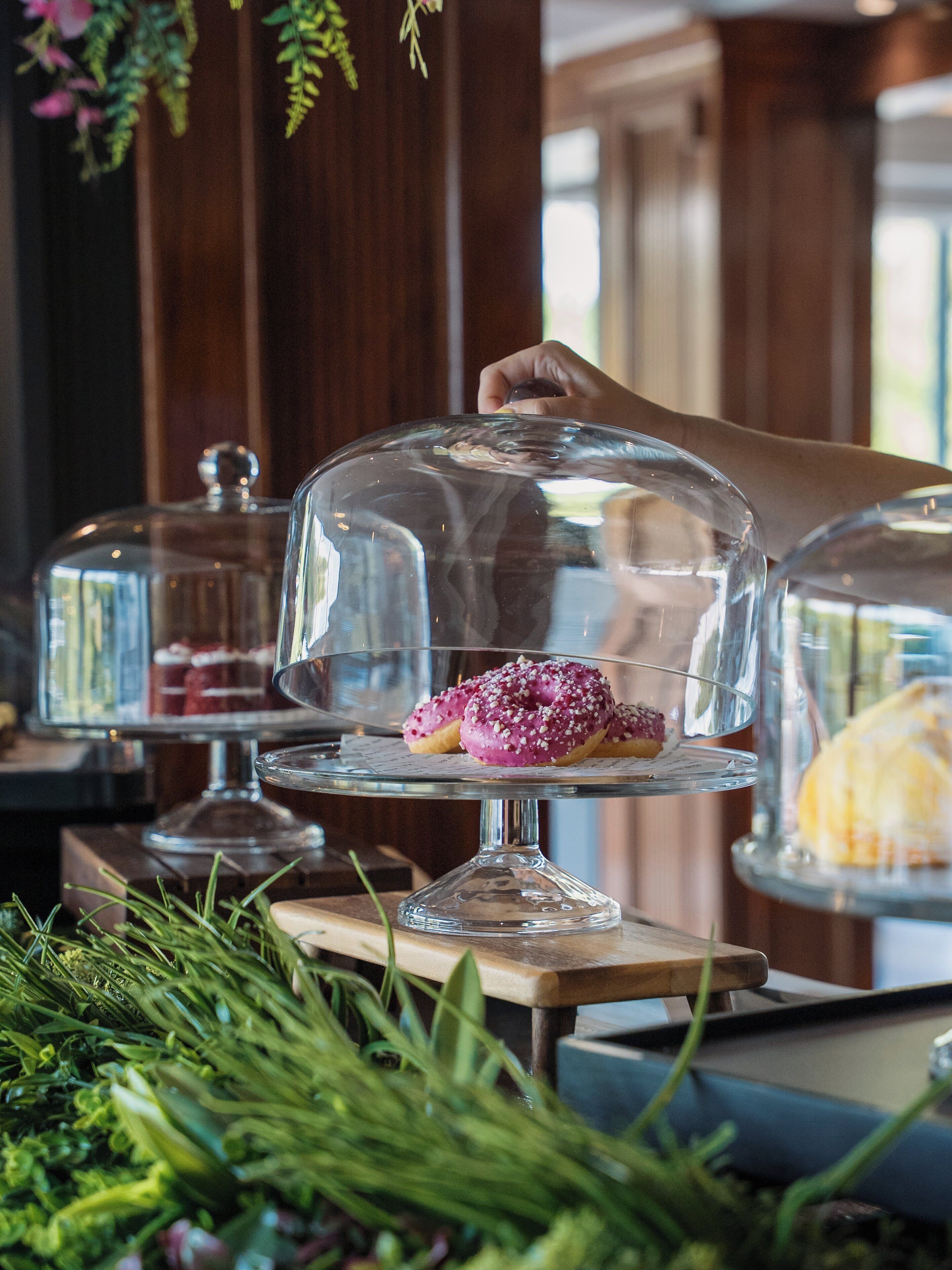 A dessert display with a hand lifting the cover, revealing pink frosted donuts with white toppings.