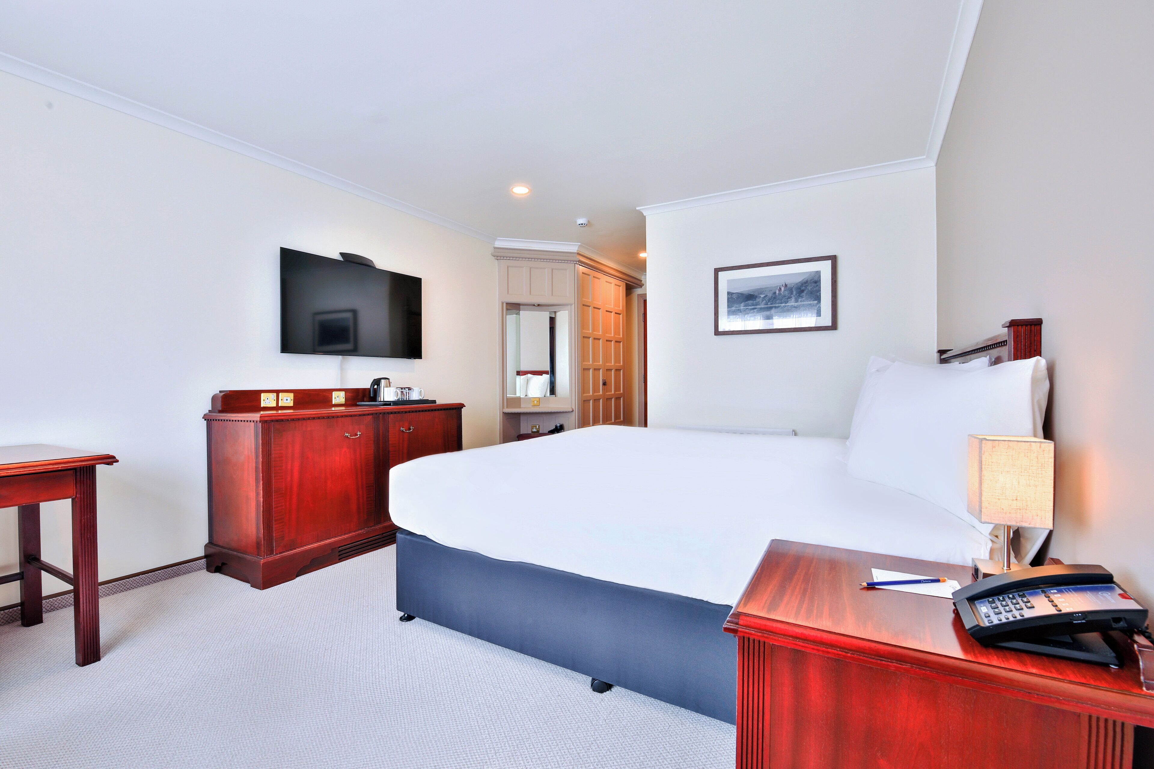 Een hotelkamer met een groot bed, een houten dressoir, een bureau met telefoon, een aan de muur bevestigde televisie en een lijst met een afbeelding.