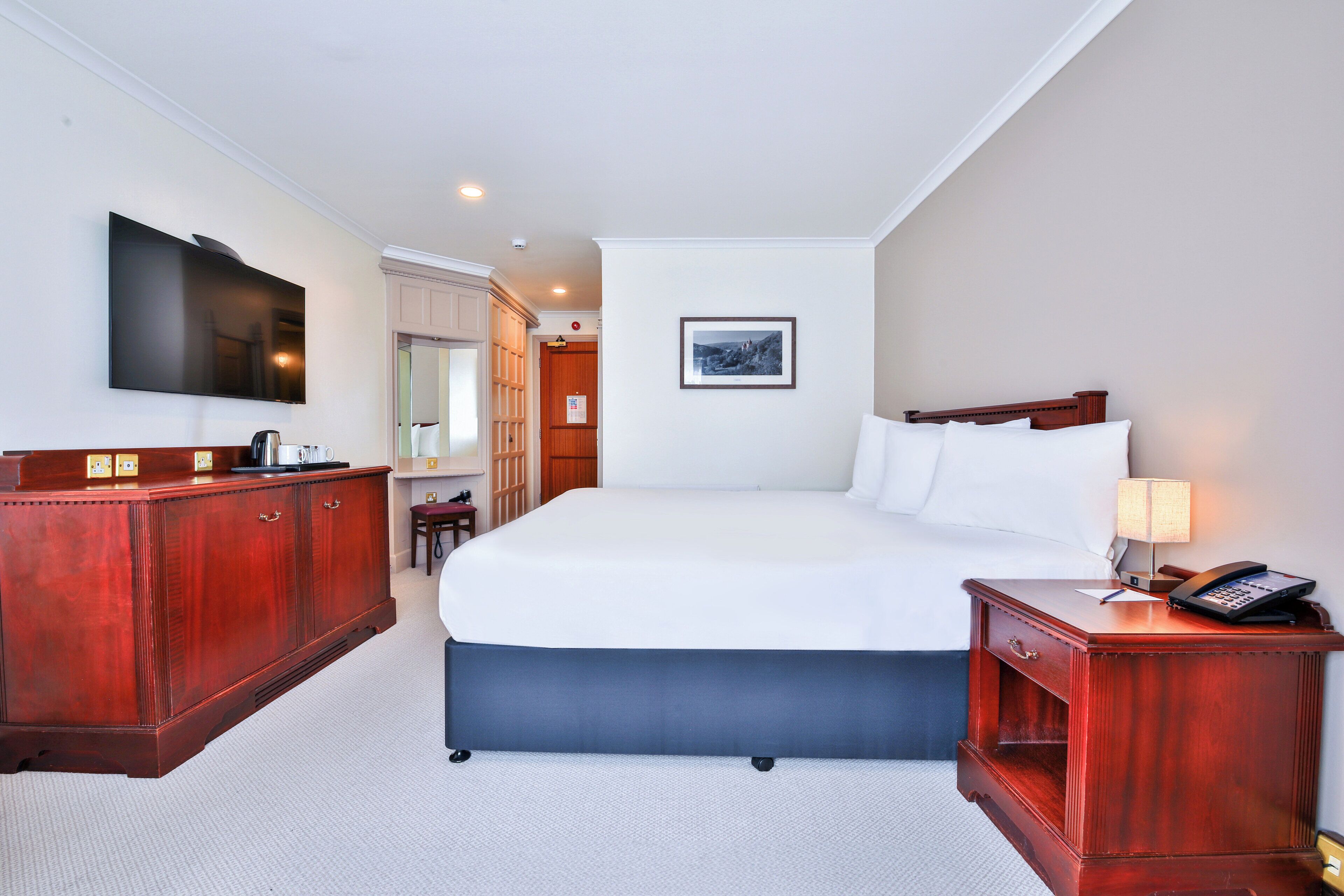 Quarto de hotel com uma cama grande, penteadeira de madeira, televisão presa na parede e um quadro enquadrado na parede.
