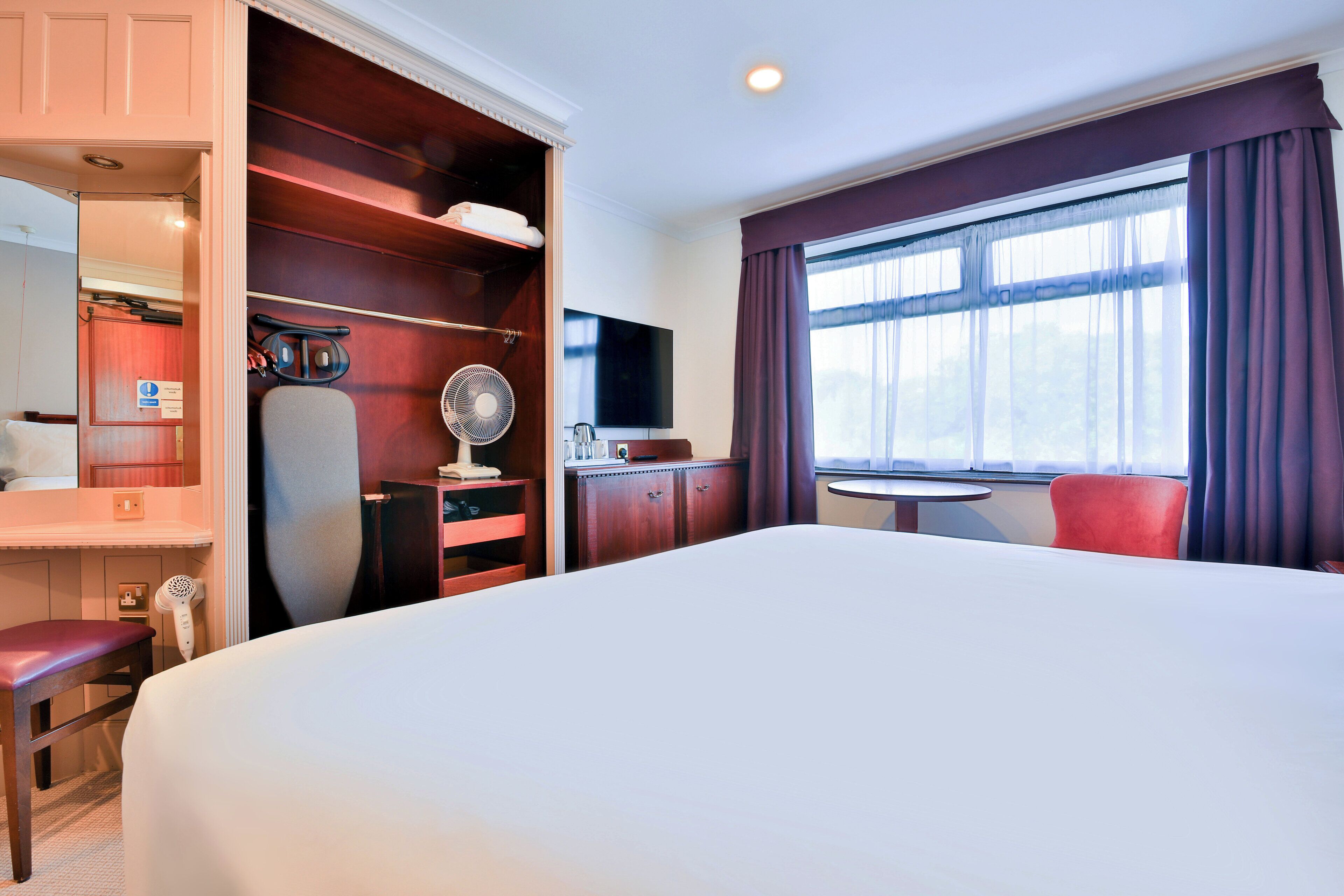 Une chambre d’hôtel avec un grand lit, un bureau avec une chaise, une petite table, un téléviseur et une fenêtre avec des rideaux.