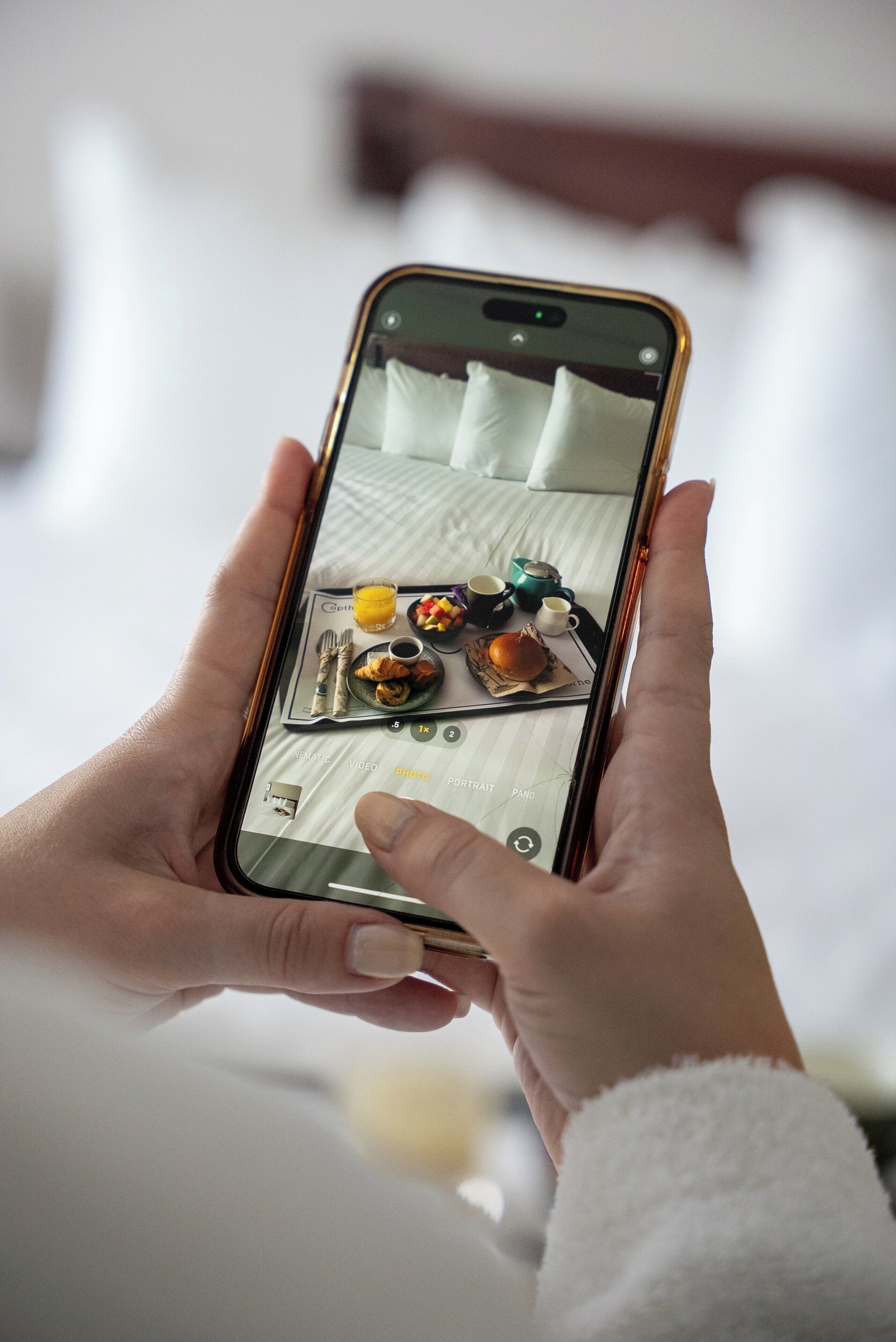 Eine Person hält ein Smartphone, auf dem ein Bett mit Kissen, eine Tasse und ein Teller mit Essen zu sehen sind.