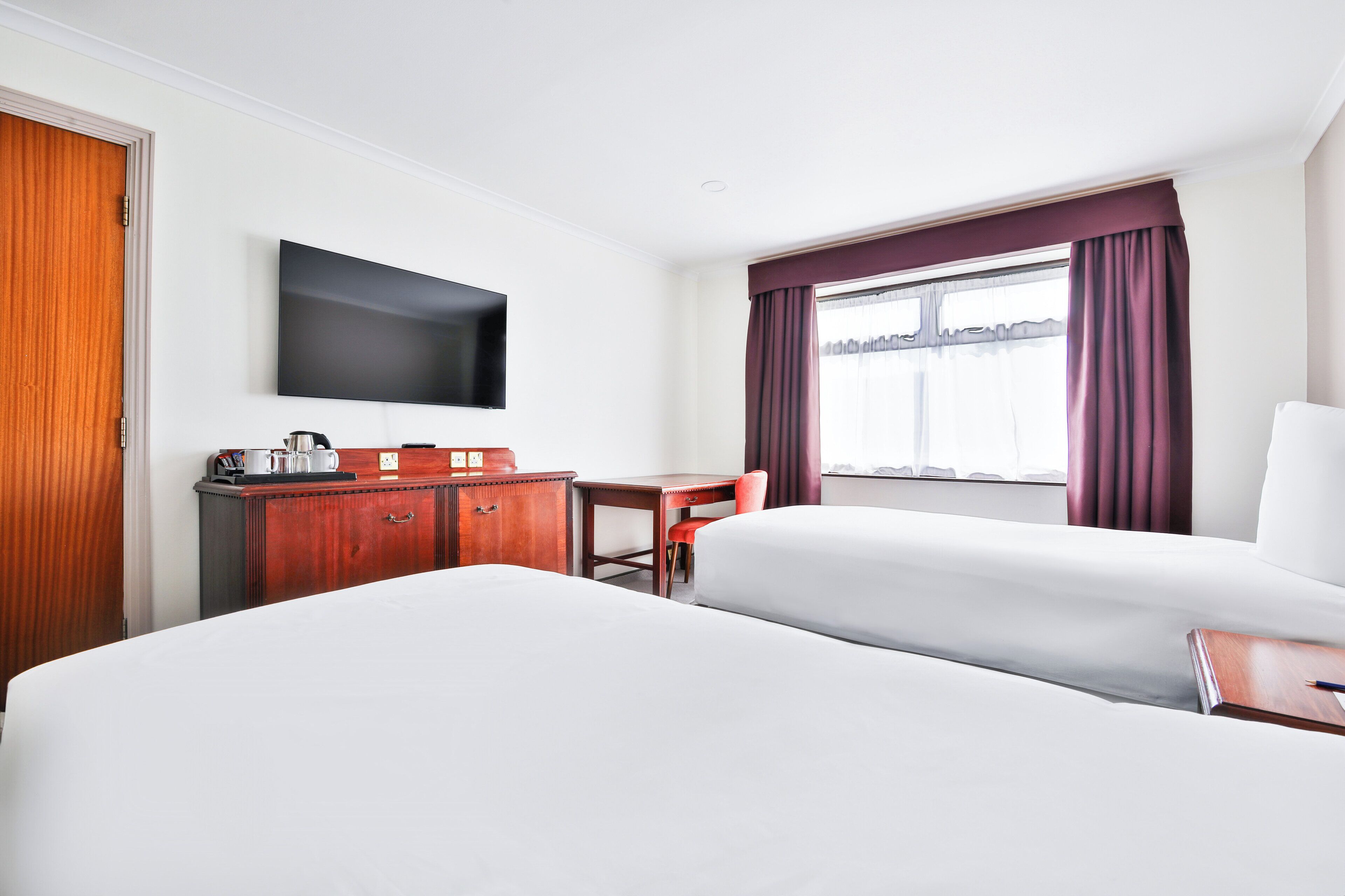 Ein Hotelzimmer mit zwei Betten, einem Schreibtisch, einem Stuhl, einem Flachbildfernseher und einem Fenster mit Vorhängen.