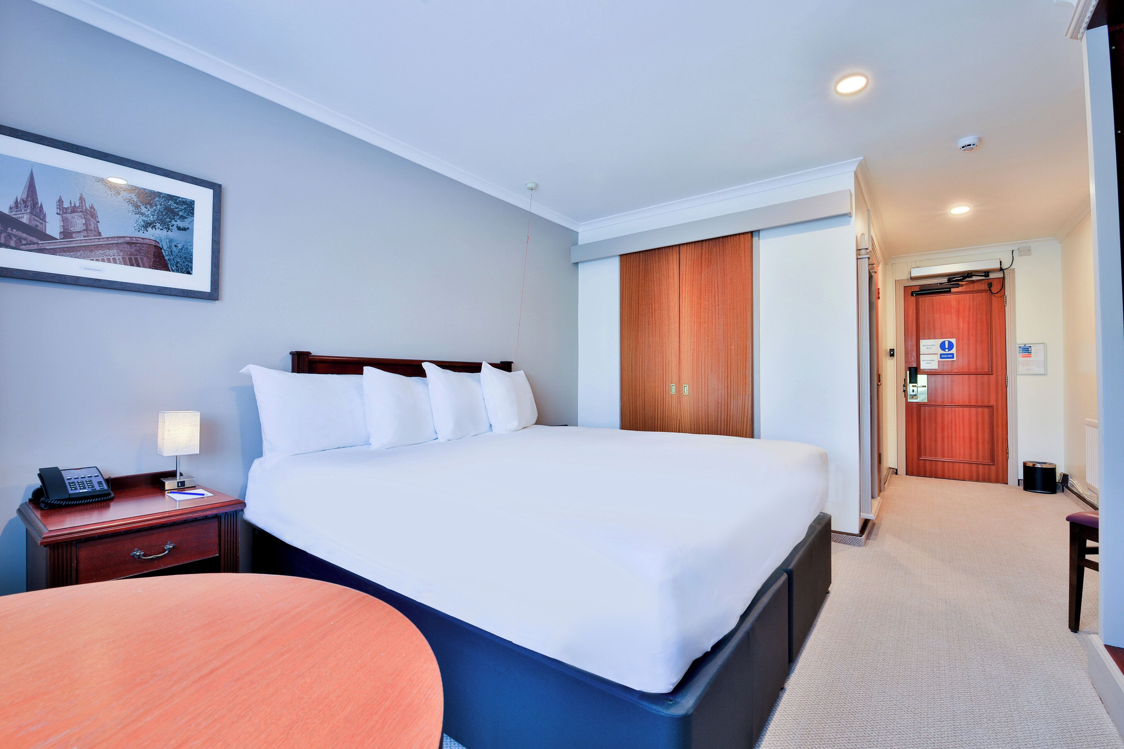 A hotel room with a large bed, a bedside table with a lamp, a wooden nightstand, and a framed picture on the wall.