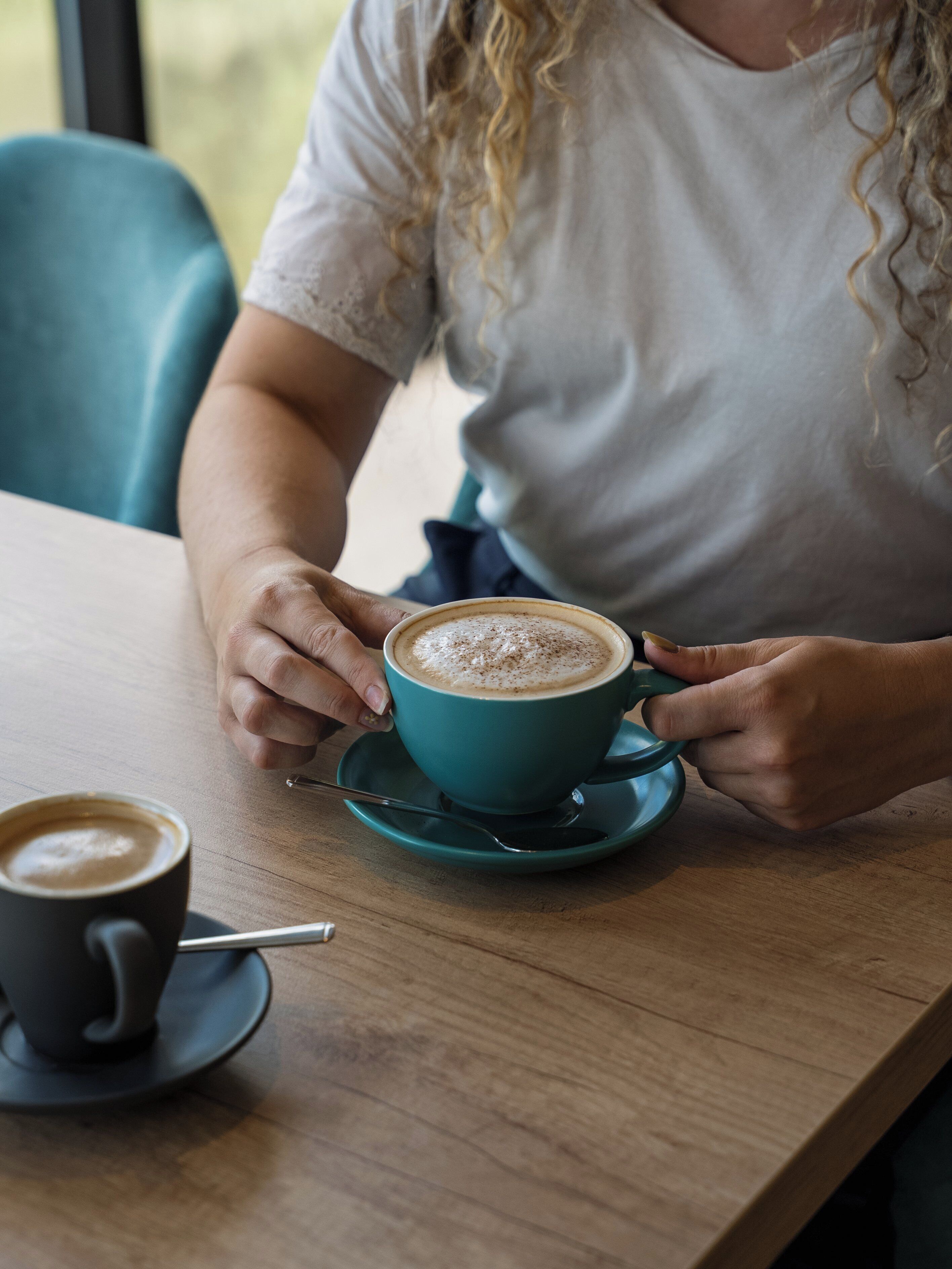 Una persona sosteniendo una taza de café con platillo sobre una mesa de madera.
