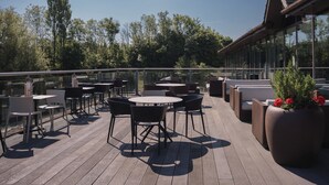 Una terrassa exterior amb taules i cadires rodones, una barana de vidre i vistes a arbres i un edifici.