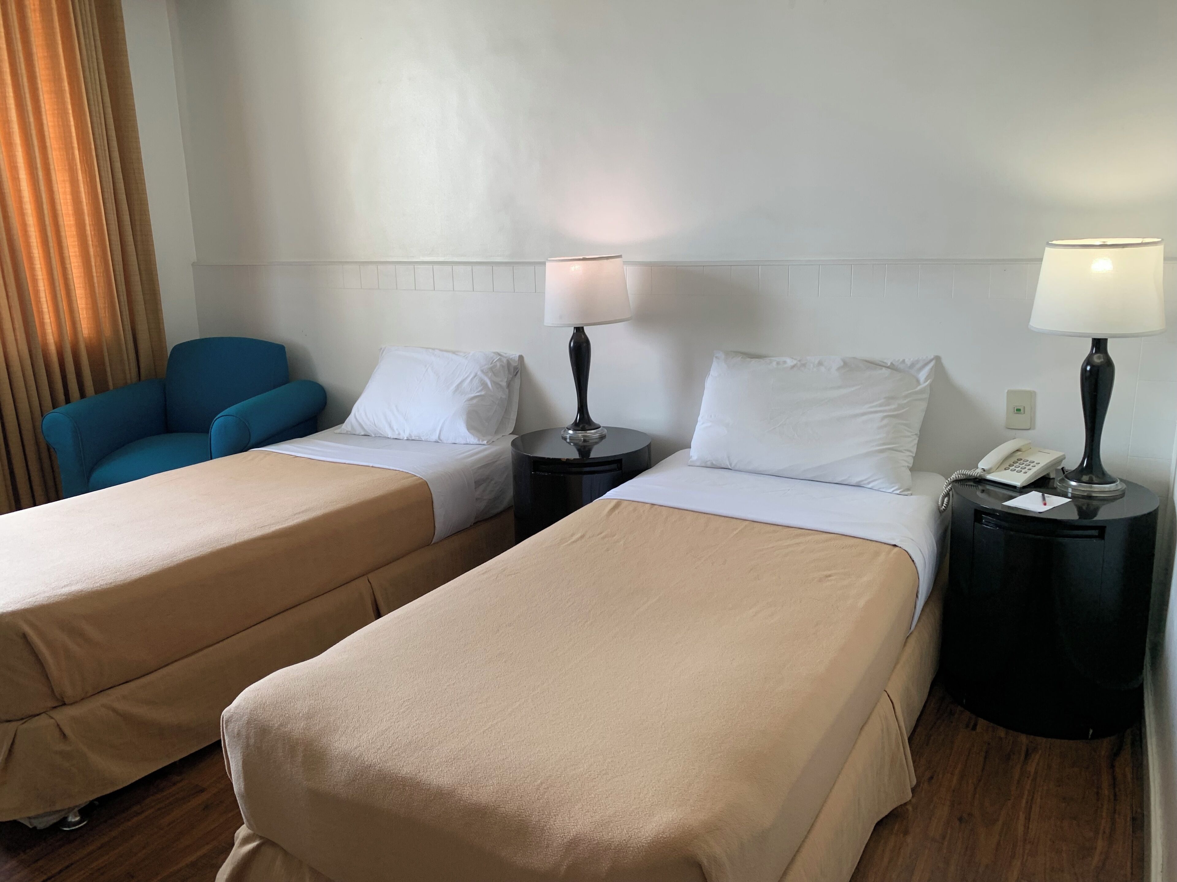 Habitación de hotel con dos camas individuales, una silla azul, una mesita de noche con lámpara y un teléfono.