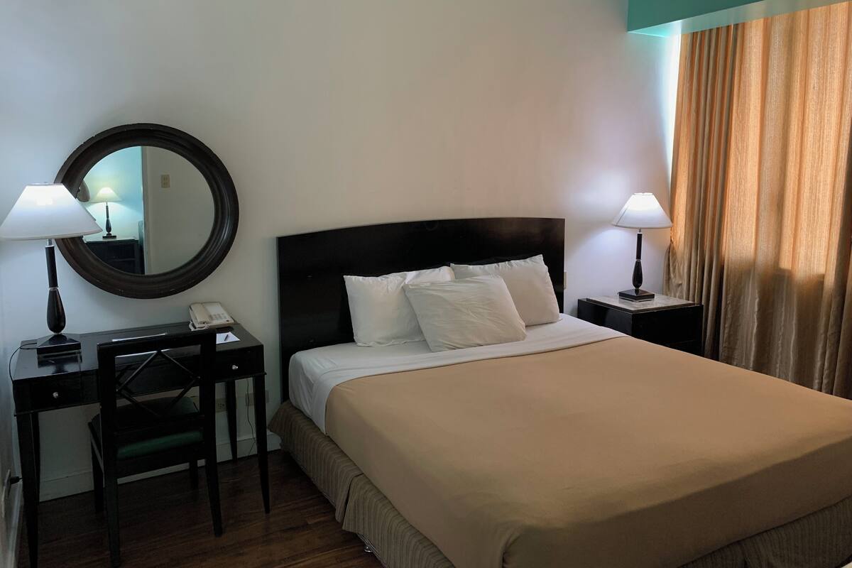 A hotel room with a bed, two bedside tables, a desk with a chair, a mirror, and a window with curtains.