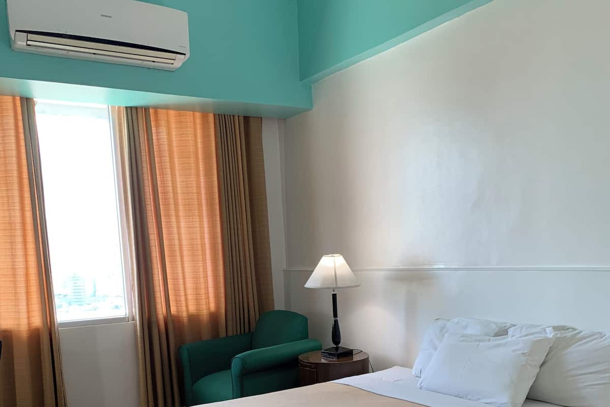 A hotel room with a large bed, a desk with a lamp, a chair, and an air conditioning unit.
