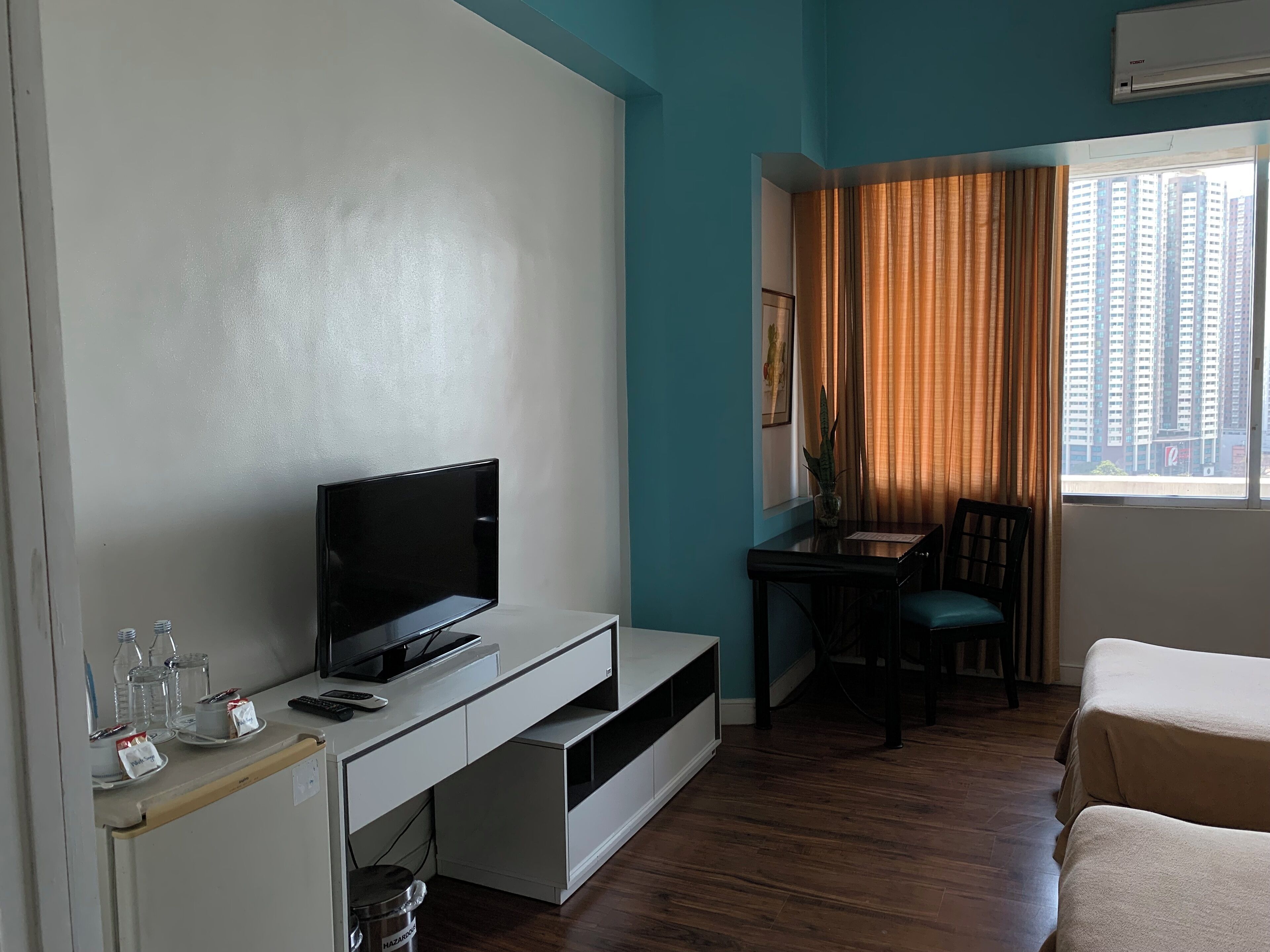 A hotel room with a flat-screen TV on a white entertainment unit, a small desk with a chair, a window with curtains, and a bed.