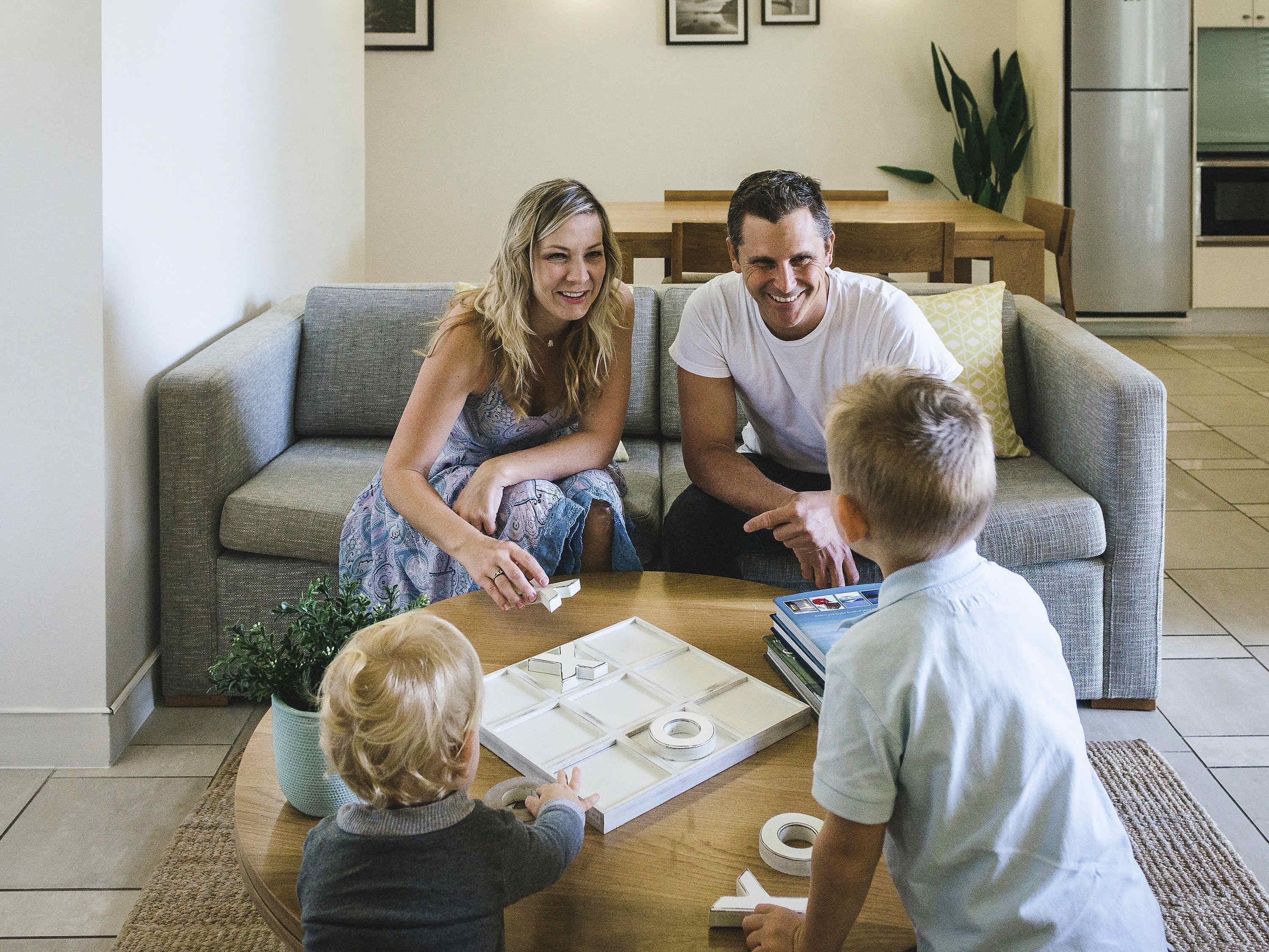 A family of three seated on a grey sofa in a living room with a coffee table, a plant, and a kitchen in the background.