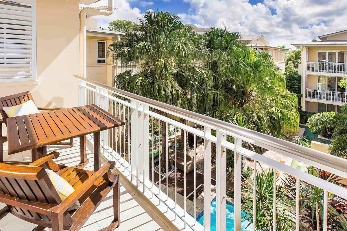 A balcony with wooden furniture, a pool, and surrounding greenery.
