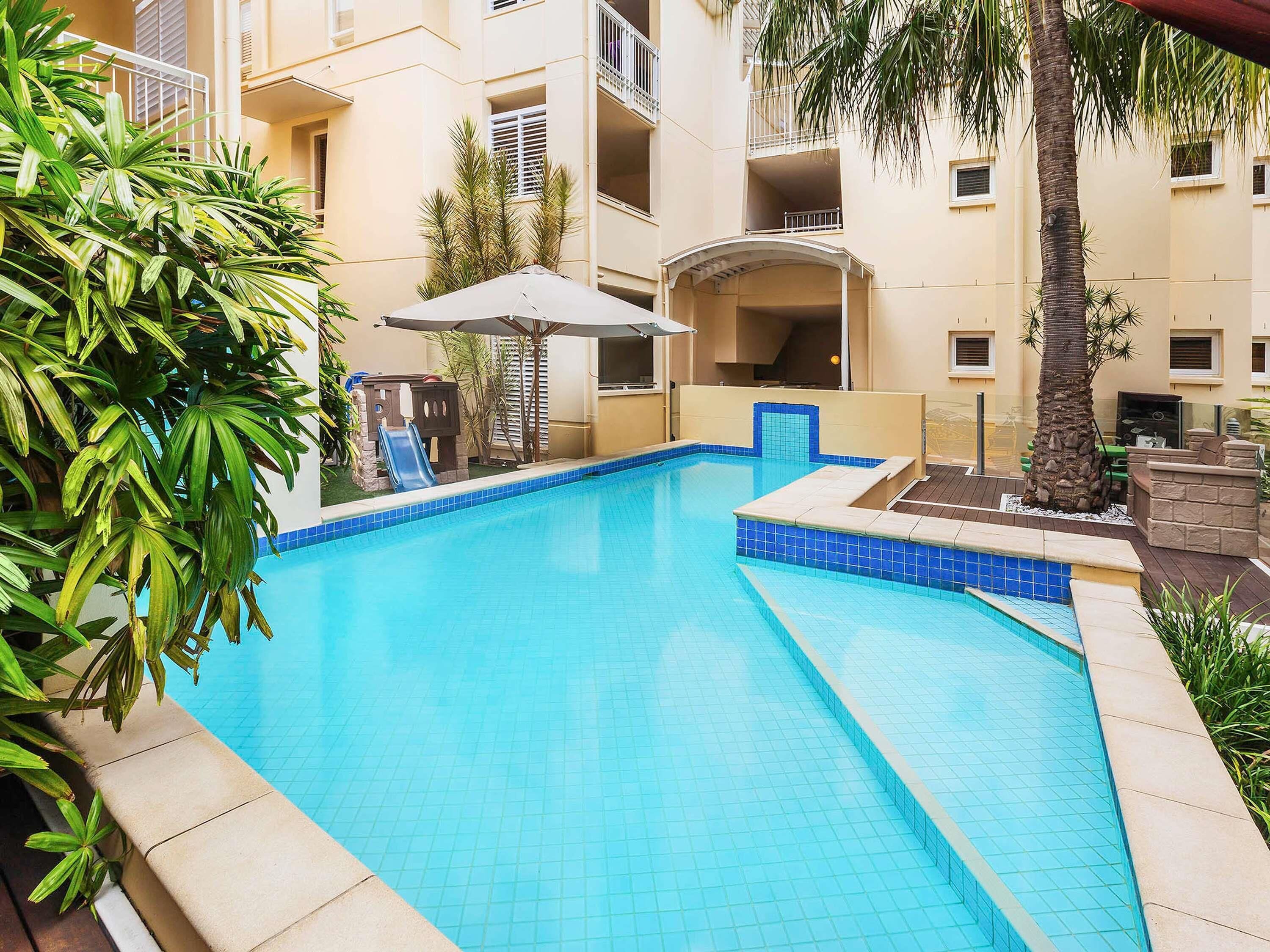 Una piscina con bordes de azulejos, rodeada por un edificio con balcones y una palmera.