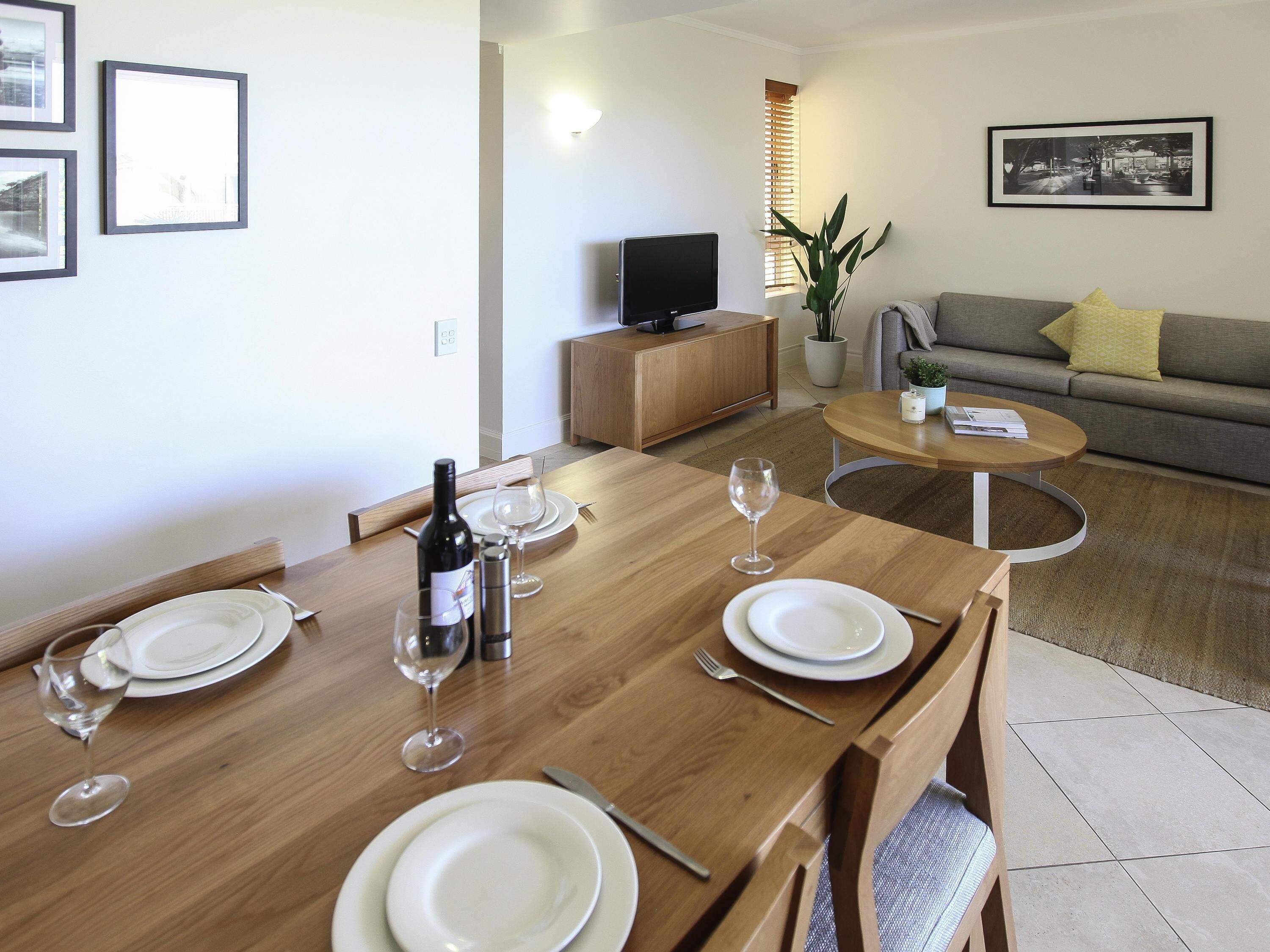 A dining area with a wooden table set for two, a television, and a sofa in the background.
