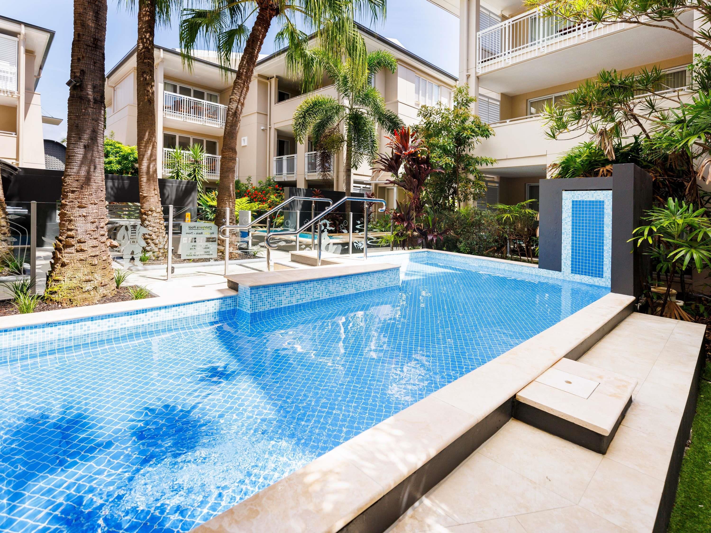 A swimming pool with tiled edges, surrounded by palm trees and a building with balconies.