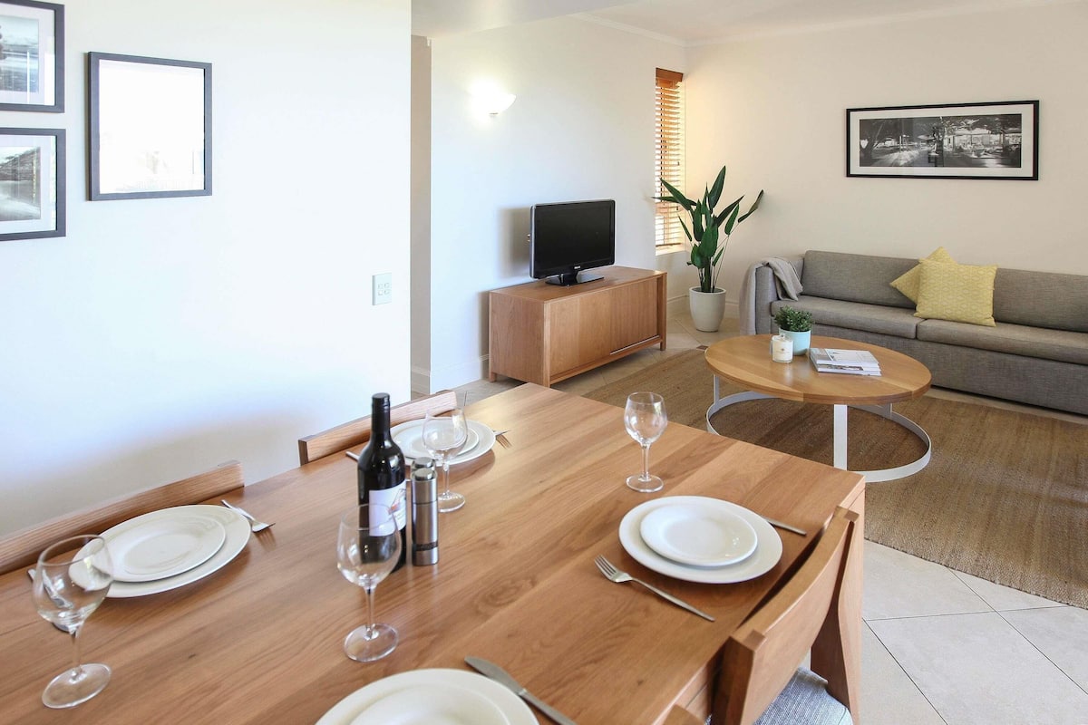 A dining area with a wooden table set for two, a television, and a sofa in the background.
