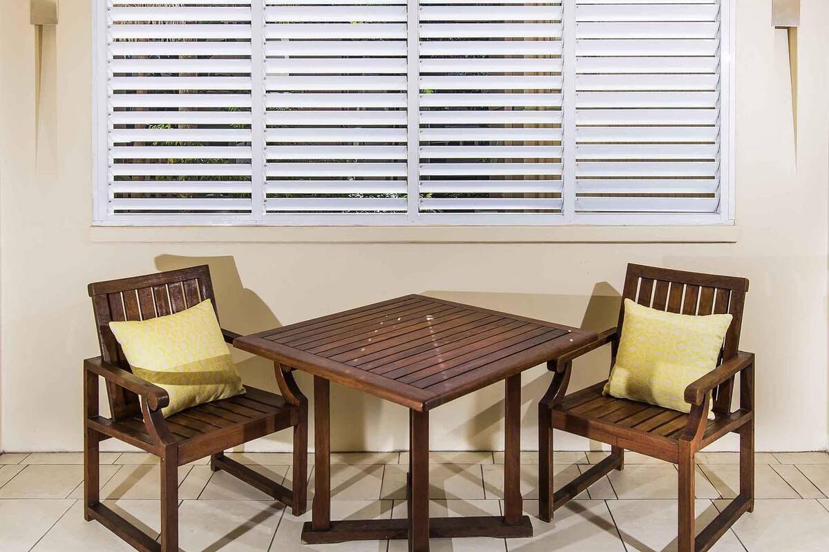 Una mesa de madera y dos sillas con cojines amarillos, ubicadas frente a una ventana con persianas blancas.