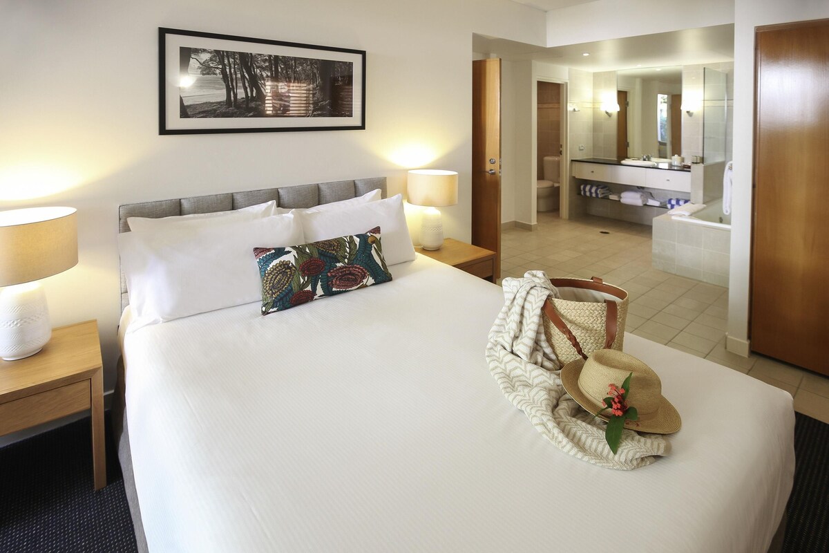 Habitación de hotel con una cama bien hecha, una lámpara de noche, un cuadro en la pared, un baño con espejo y lavamanos, y una puerta que conduce a otra habitación.