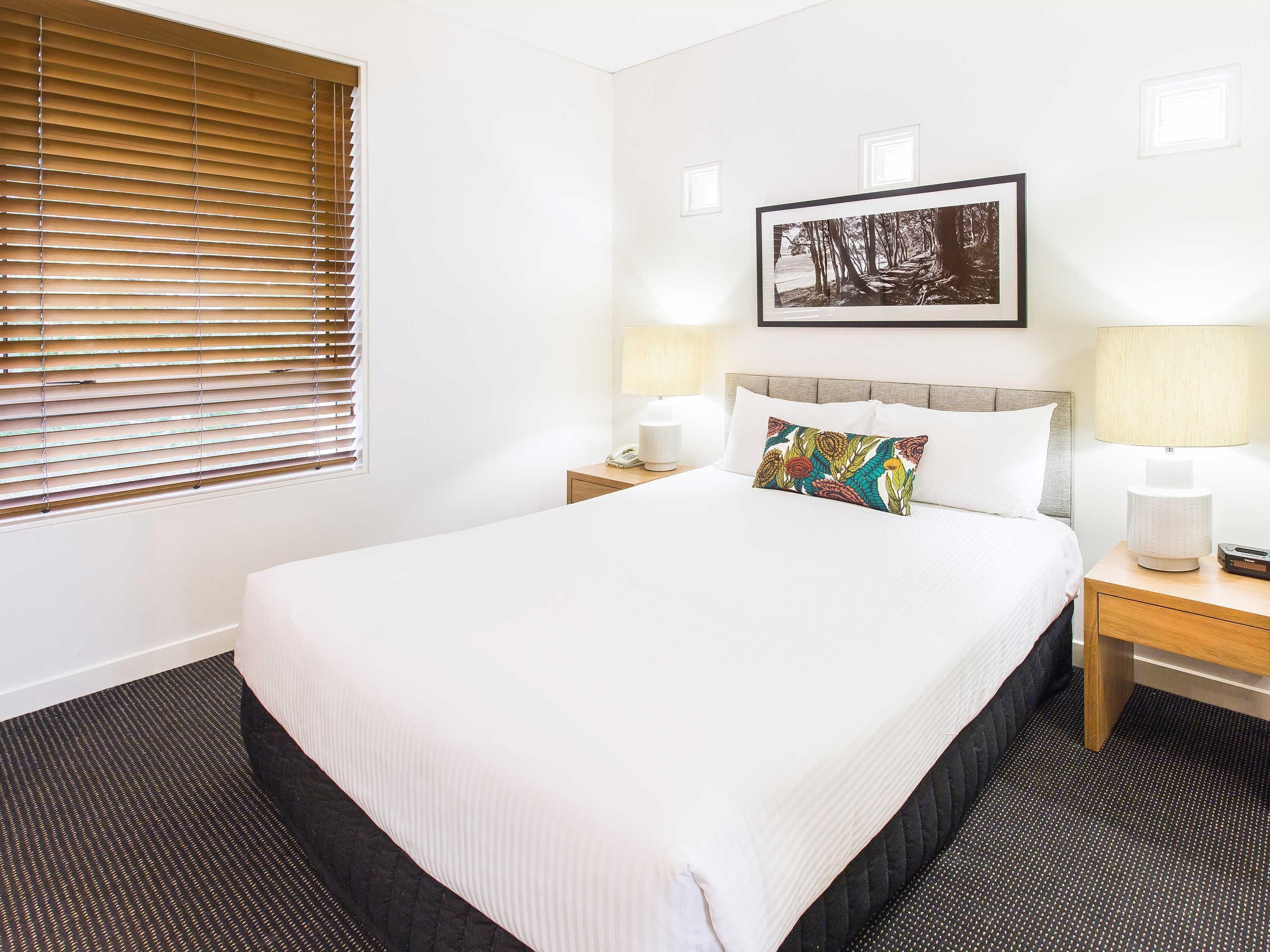 A hotel room with a large bed, a bedside table with a lamp, a framed picture on the wall, and a window with blinds.