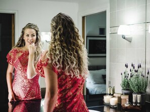 Eine Frau im roten Kleid steht vor einem Spiegel in einem Badezimmer, auf der Ablage stehen ein Topf mit einer Pflanze und Kerzen.