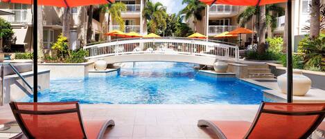 A pool area with lounge chairs and umbrellas, a bridge, and a building with balconies.