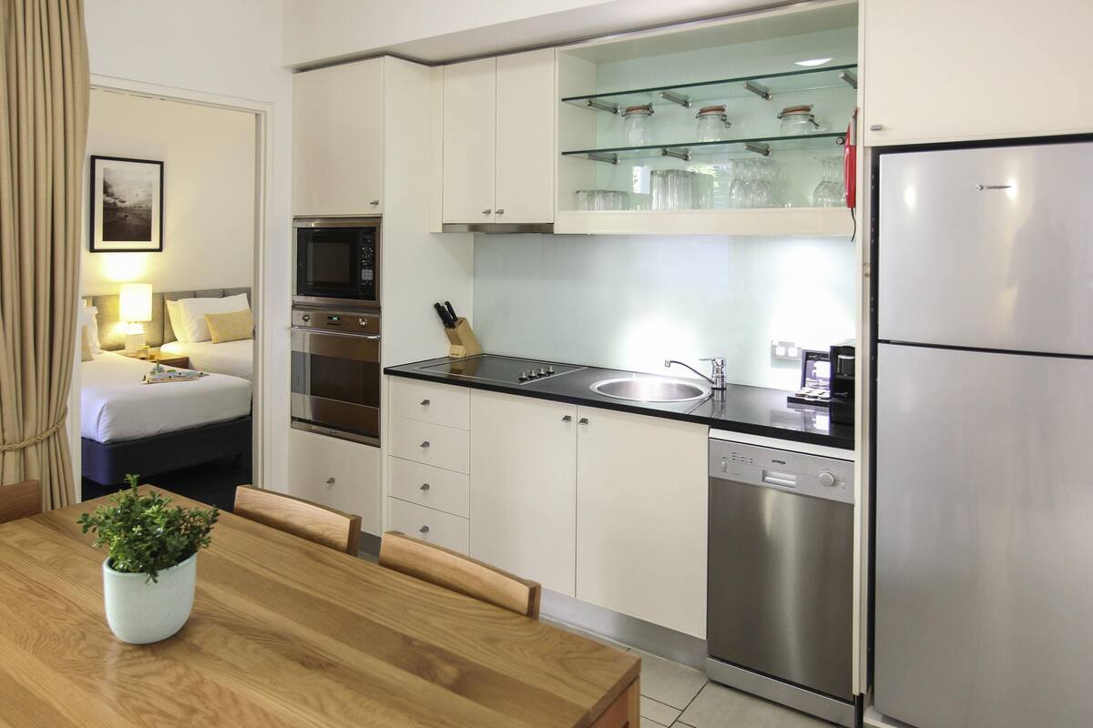 A modern kitchen with stainless steel appliances, a glass-fronted cabinet with jars, a dining area with a wooden table and chairs, and a bedroom with a bed and nightstand.