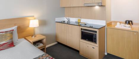 Habitación de hotel pequeña y funcional, con cama, mesita de noche con lámpara, y una kitchenette con estufa, microondas y pileta.