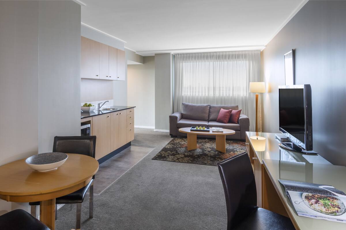 A modern hotel room with a kitchenette, a dining area, a sofa, and a TV.