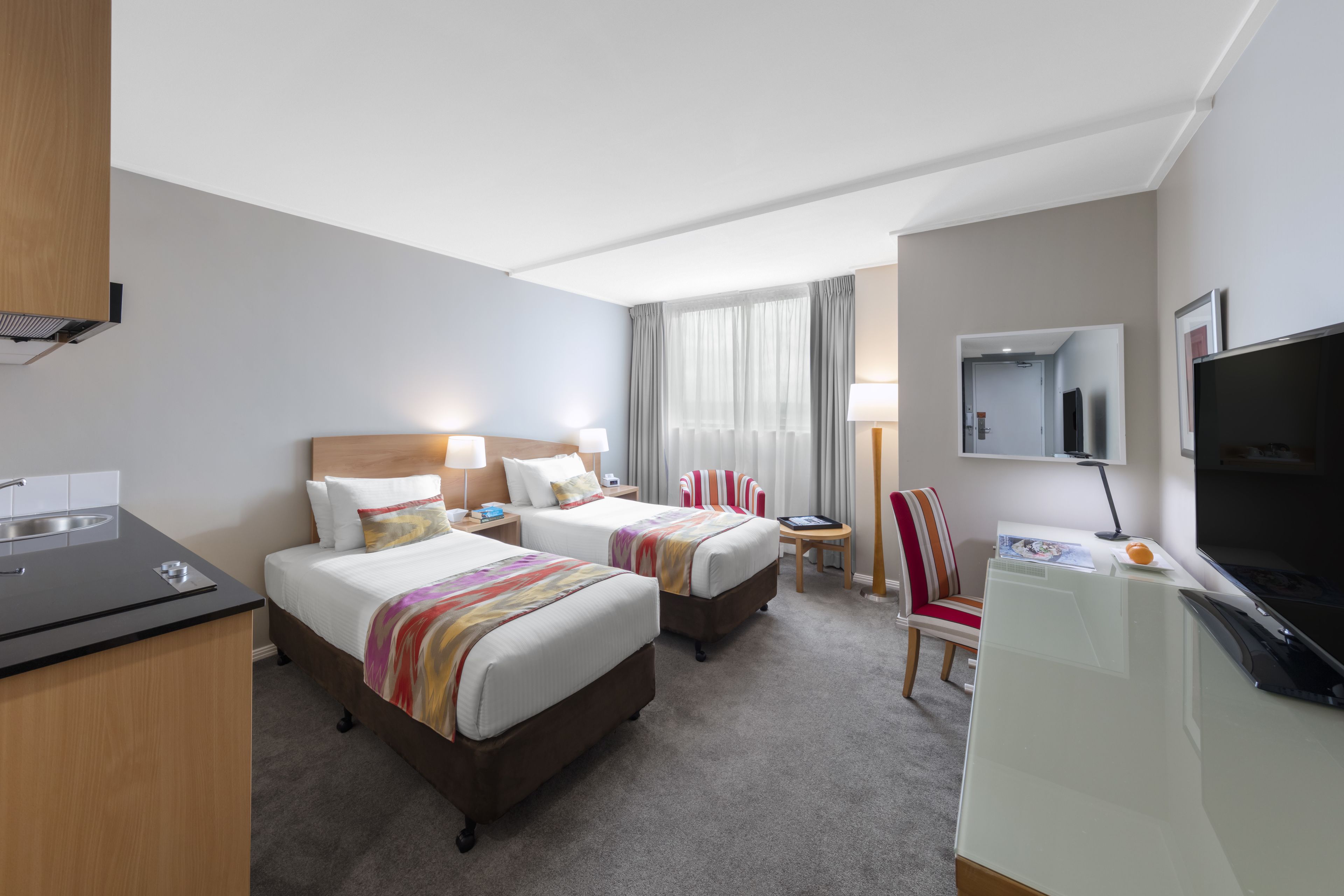 A hotel room with two beds, a desk, a TV, and a small table with a lamp.