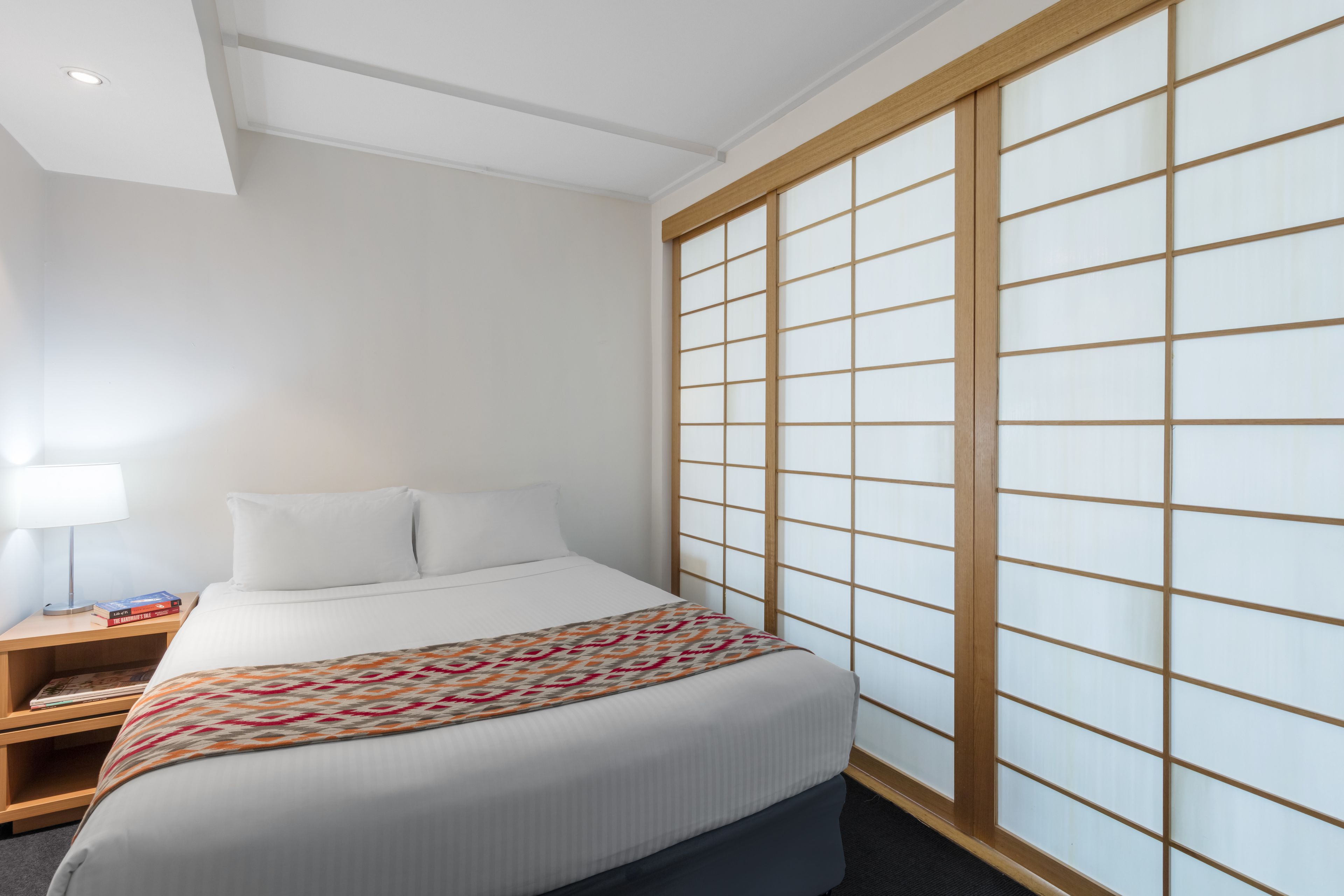 Une chambre d’hôtel avec un lit, une table de chevet avec des livres, une lampe et un paravent japonais.