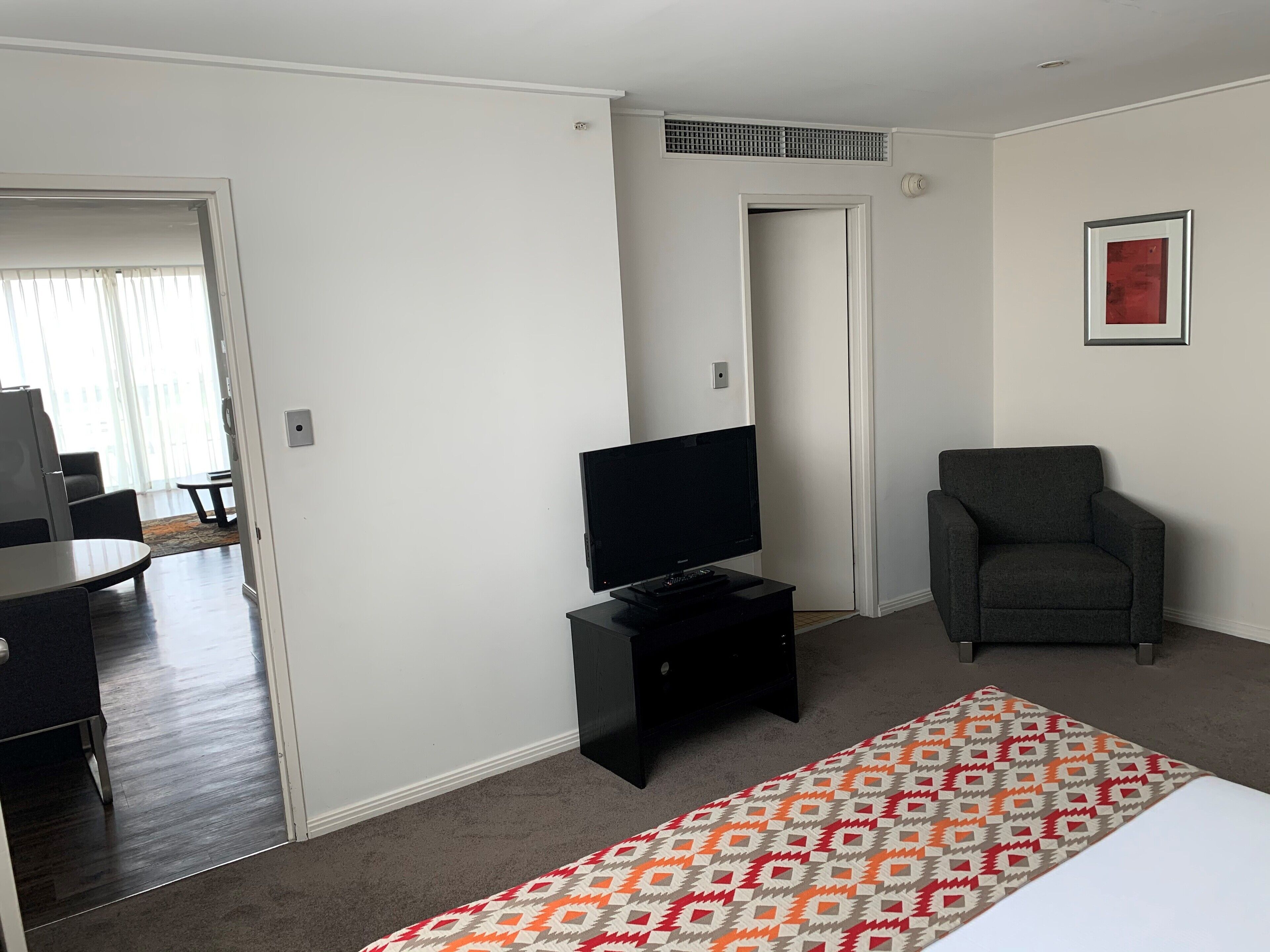 A hotel room with a bed, a TV on a stand, a grey armchair, and a patterned rug.