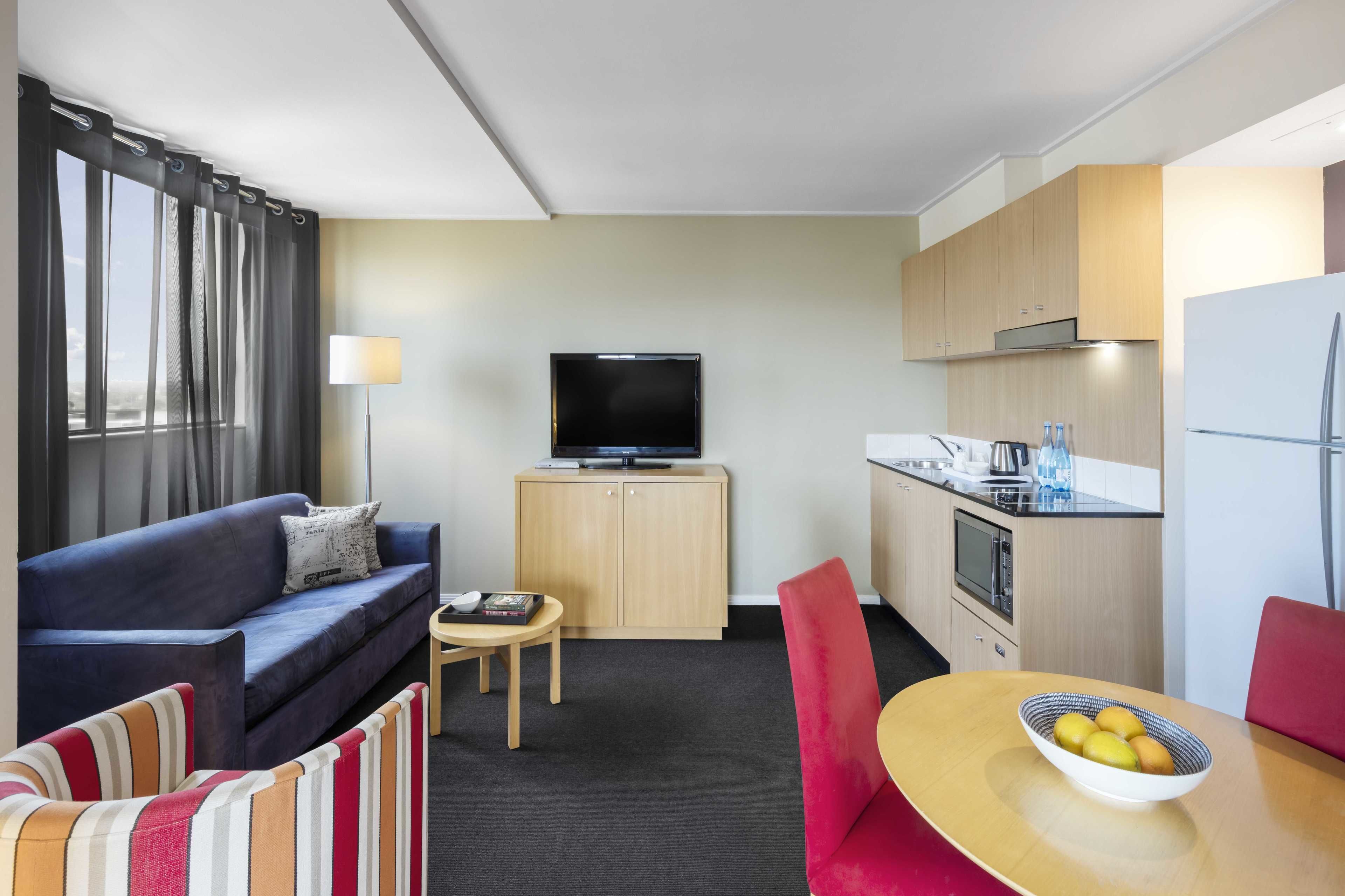 Habitación de hotel compacta con sofá, una mesita, televisor, zona de cocina y una mesa de comedor con un bol de fruta.