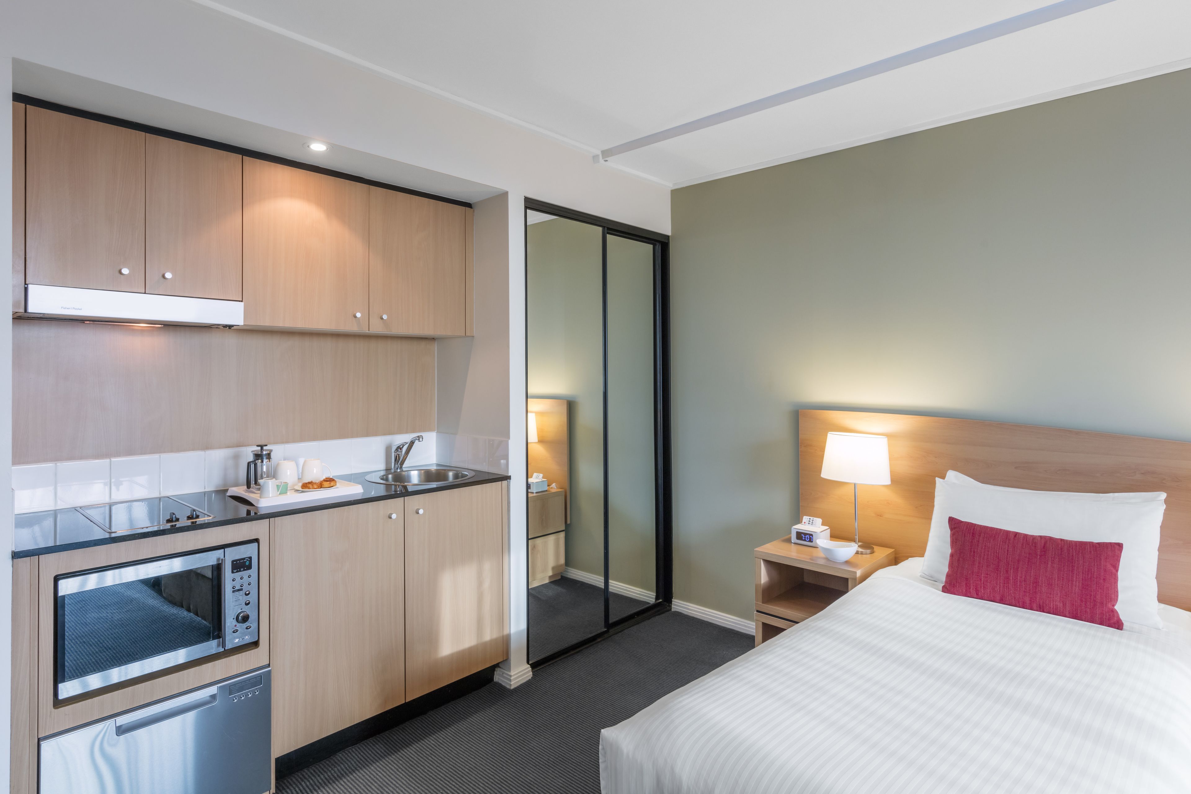 A hotel room with a kitchenette, a bed with white and red bedding, a nightstand with a lamp, and a mirror.