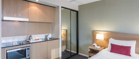 A hotel room with a kitchenette, a bed with white and red bedding, a nightstand with a lamp, and a mirror.
