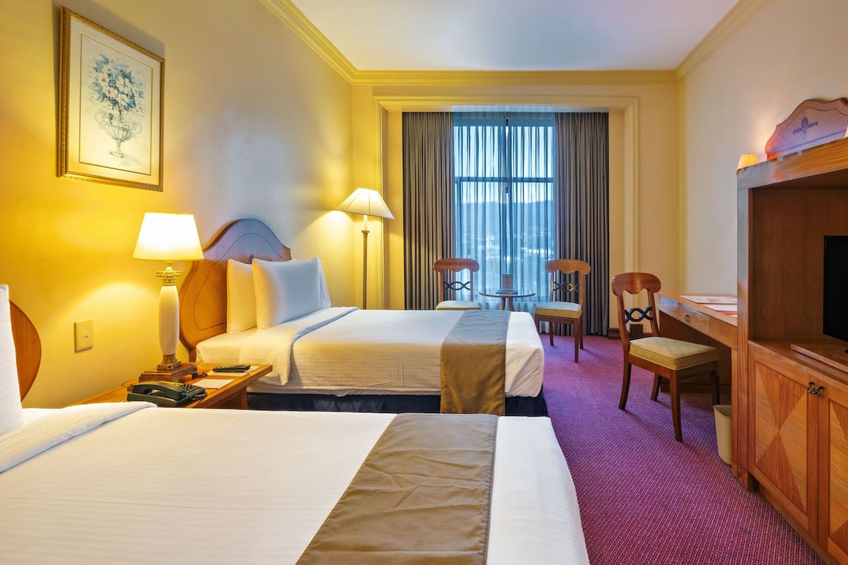 Une chambre d’hôtel avec deux lits, un bureau, une chaise, une télévision et une fenêtre avec des rideaux.