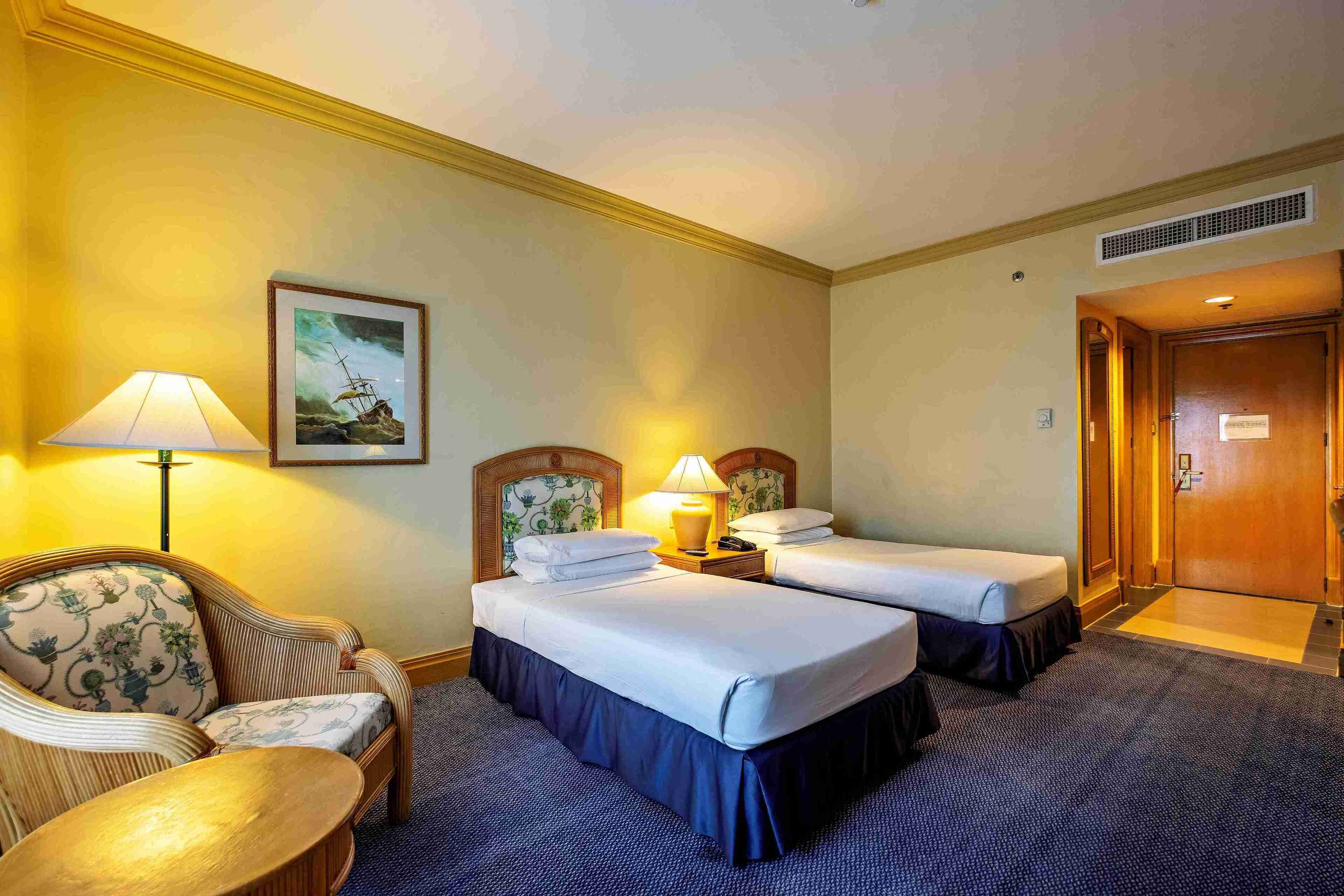 A hotel room with two beds, a chair, a lamp, and a painting on the wall.